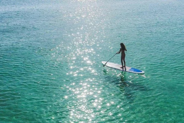 A woman paddleboarding on calm, clear ocean water with sunlight reflections.