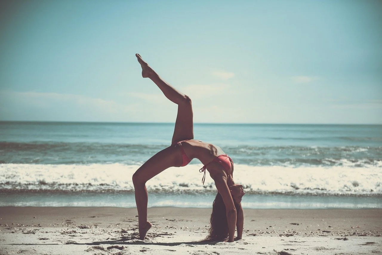 Woman in a red bikini performing a backbend yoga pose on the beach near ocean waves.