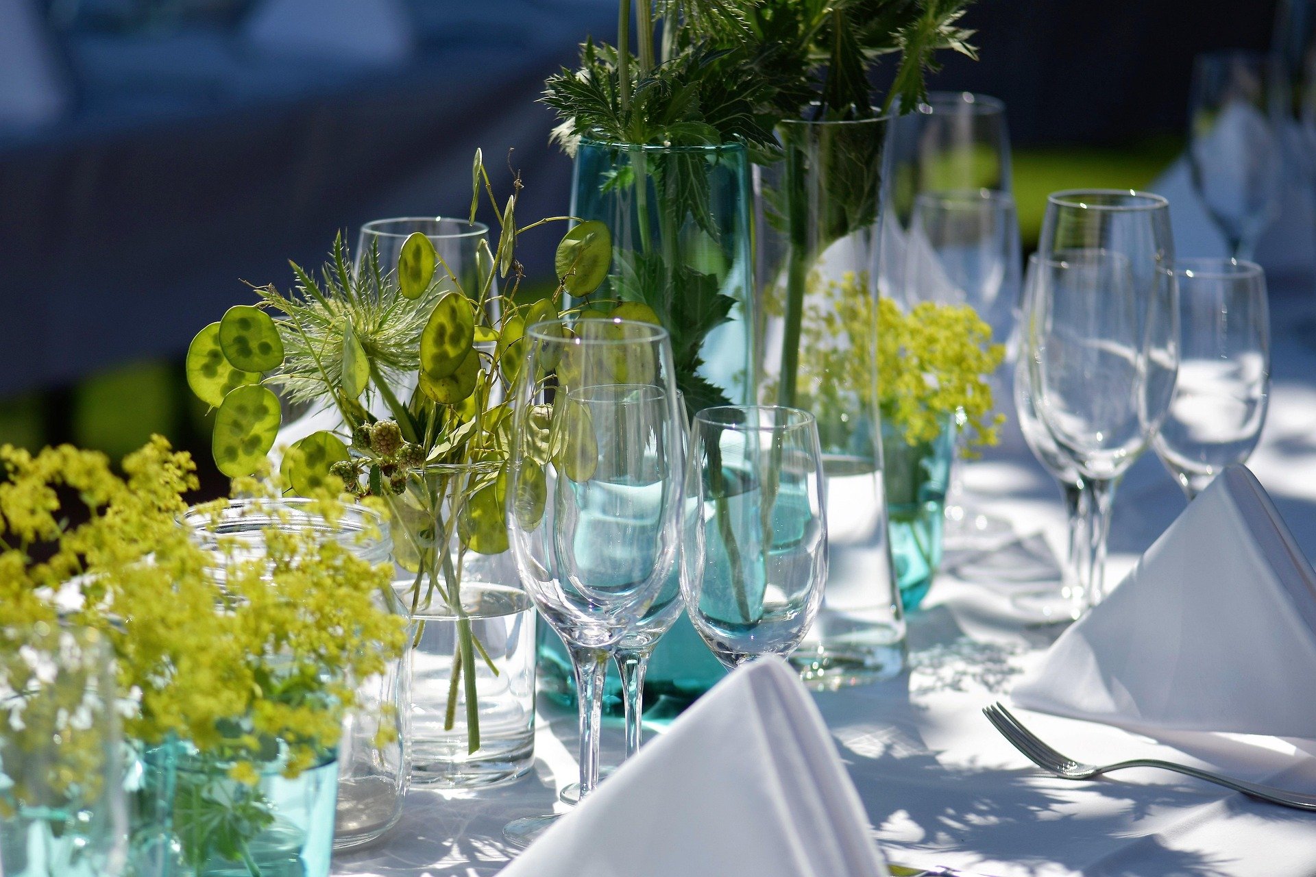 Elegant table setting with clear glass vases containing green foliage and yellow flowers, white tablecloth, white napkins, and empty wine glasses arranged outdoors.
