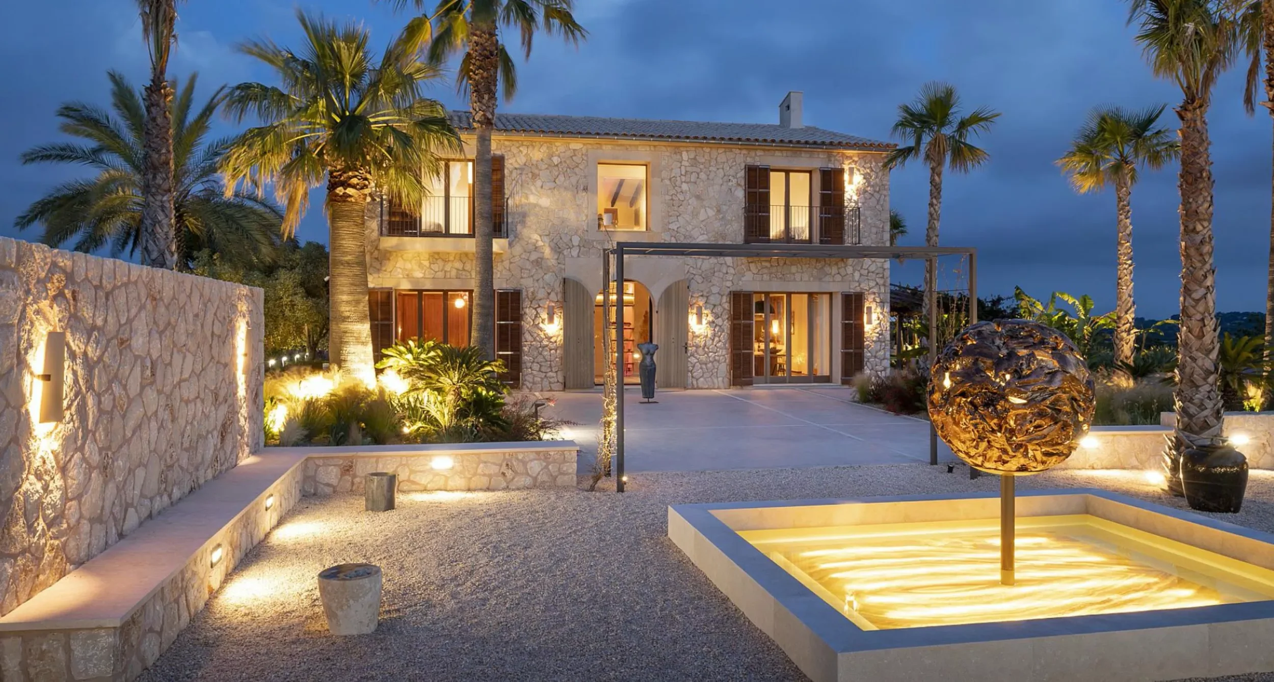 A luxurious stone house with large windows and wooden shutters, surrounded by palm trees, with a courtyard featuring a lit stone fountain, decorative lighting, and landscaping at dusk.