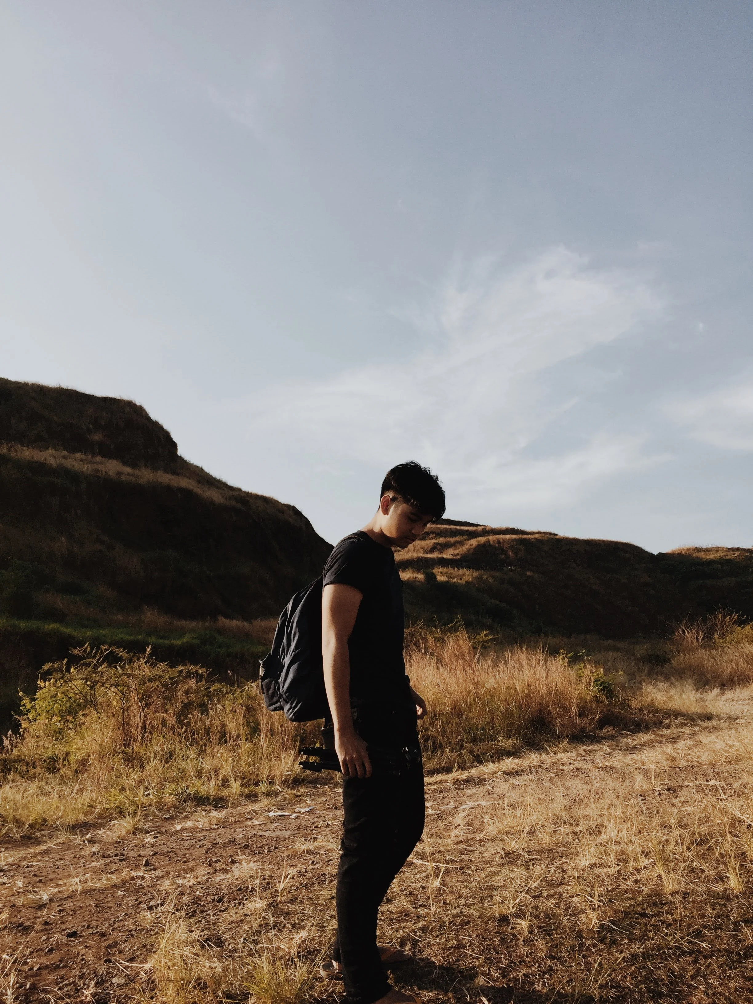 A young man stands in a dry, grassy field with rolling hills and a partly cloudy sky in the background. He is wearing a black t-shirt, black pants, and has a backpack, looking downward with a contemplative expression.