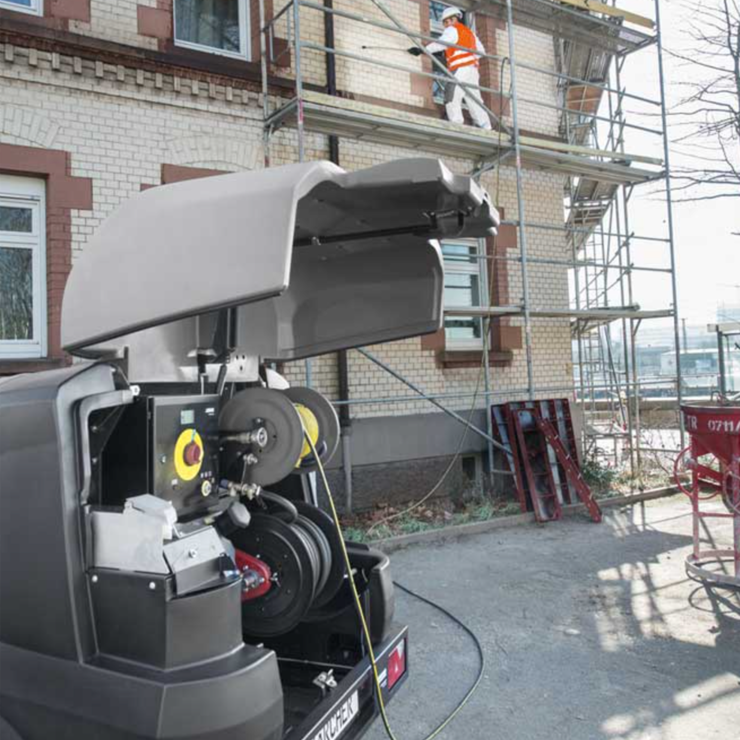 Byggearbeidere på stillas foran et mursteinsbygg, med en maskin i forgrunnen.