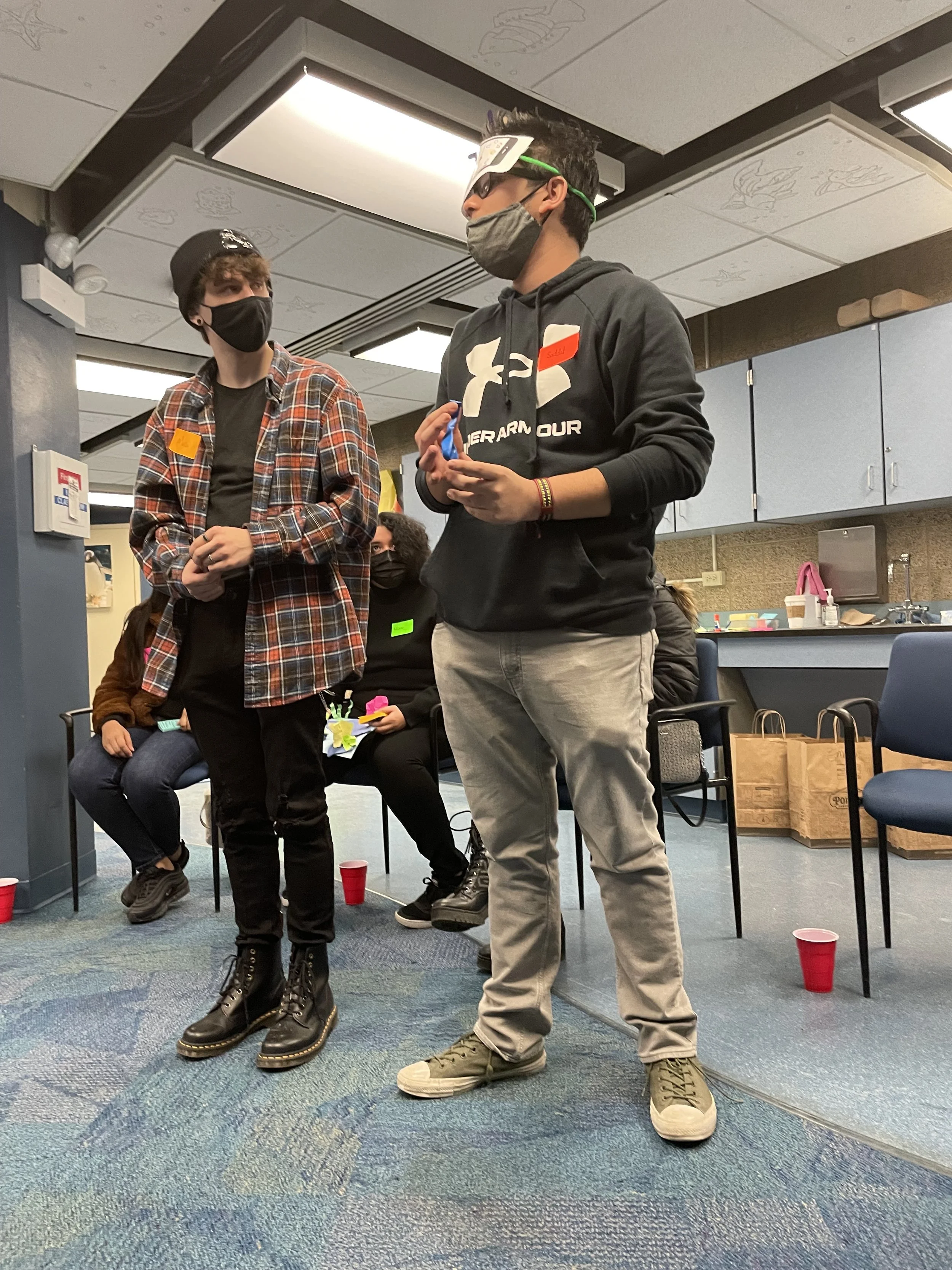 Two young men standing indoors, wearing masks and casual clothing, with other seated individuals and red plastic cups on the floor in the background.