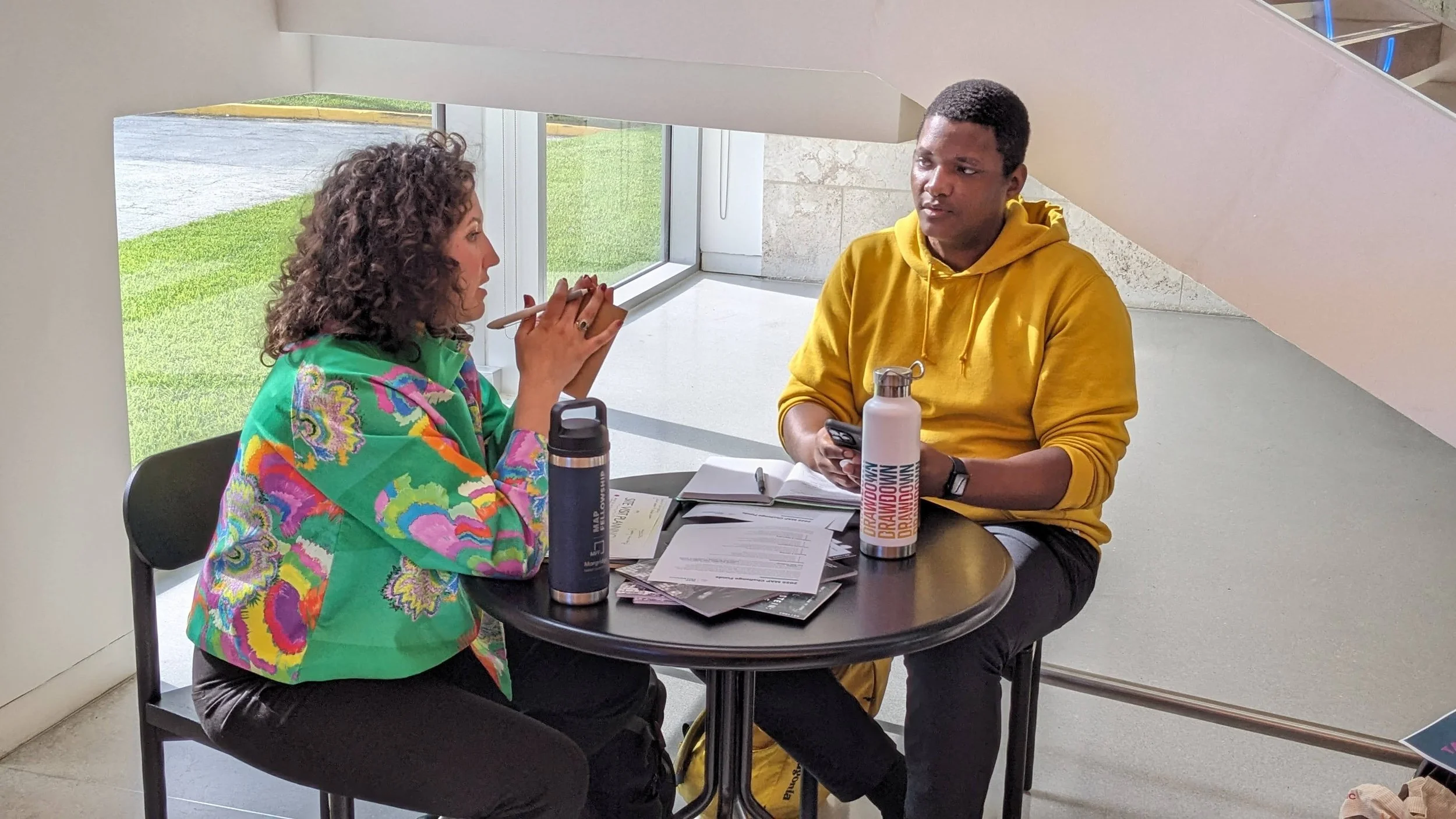 A woman and man sitting at a small round table indoors, engaged in conversation. The woman is wearing a colorful green and pink jacket, and the man is wearing a yellow hoodie. The table has papers, notebooks, and water bottles.