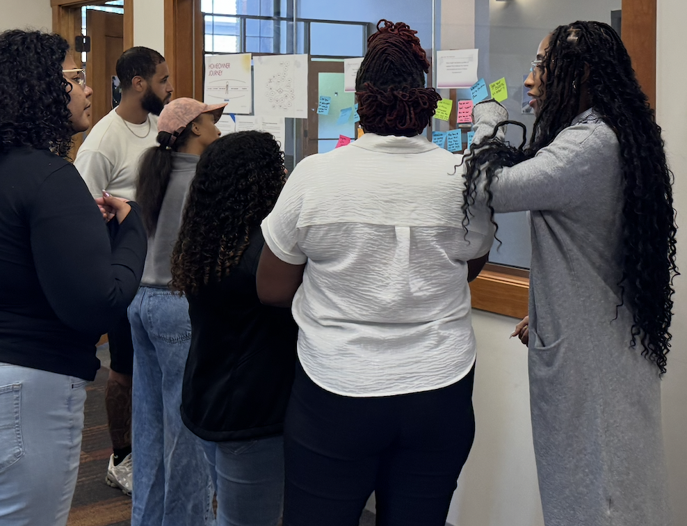 Group of seven diverse people gathered around a wall covered with colorful sticky notes and papers, engaging in a discussion or brainstorming session.
