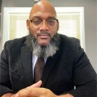 A man with glasses and a beard, dressed in a dark suit and tie, sitting indoors with a window in the background.