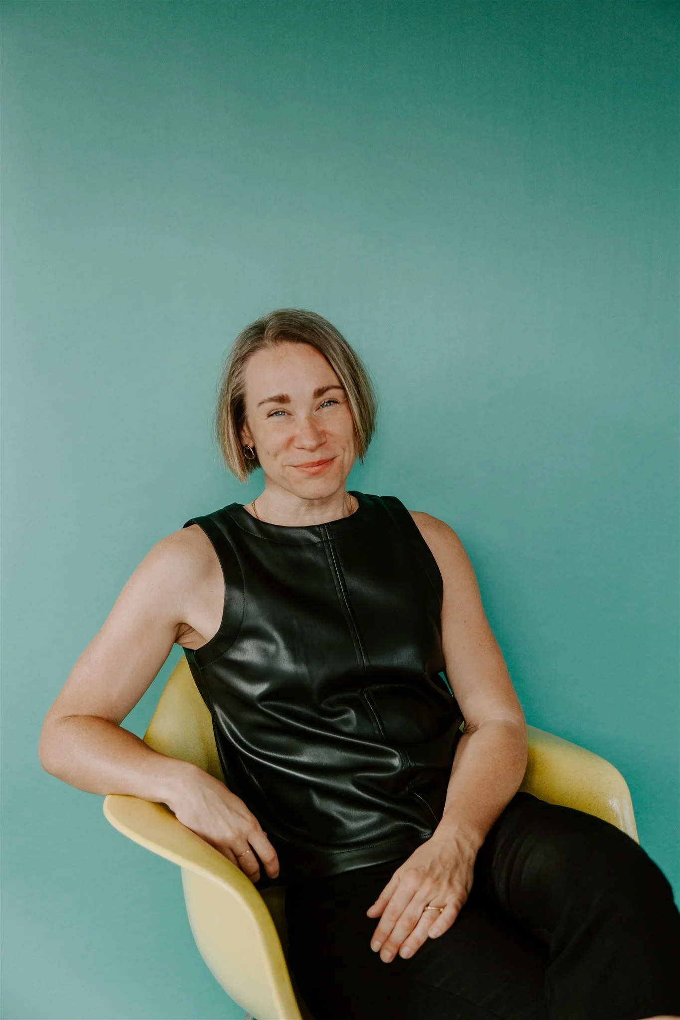 A woman with short blonde hair is sitting in a yellow chair against a teal background, wearing a sleeveless black leather top and black pants, smiling subtly.