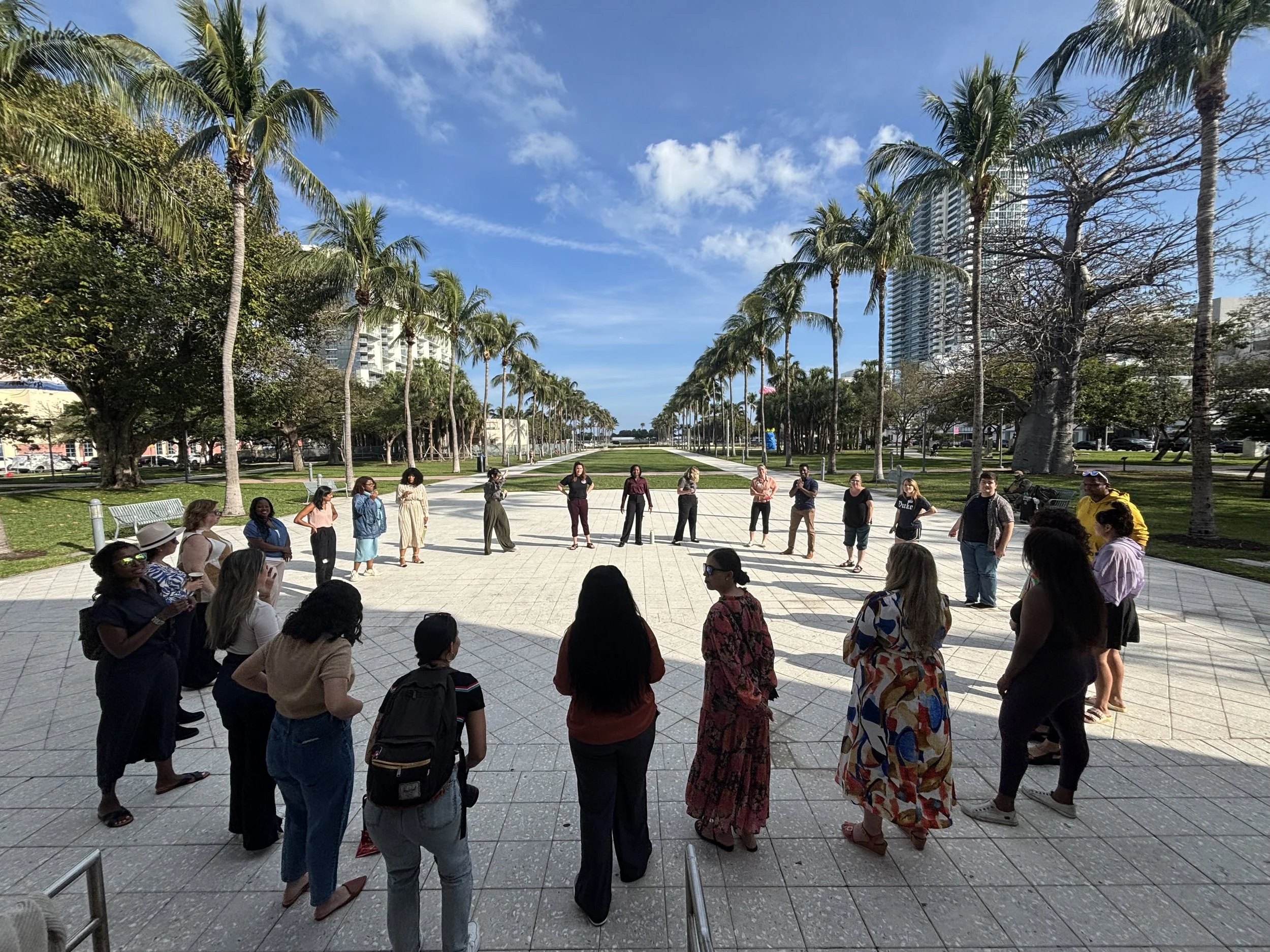 People gathered in a circle outdoors in a park with palm trees, high-rise buildings, and a blue sky in the background.