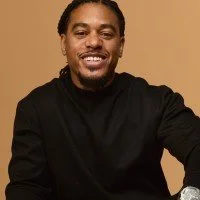 A smiling man with long braided hair, wearing a black shirt, against a neutral background.