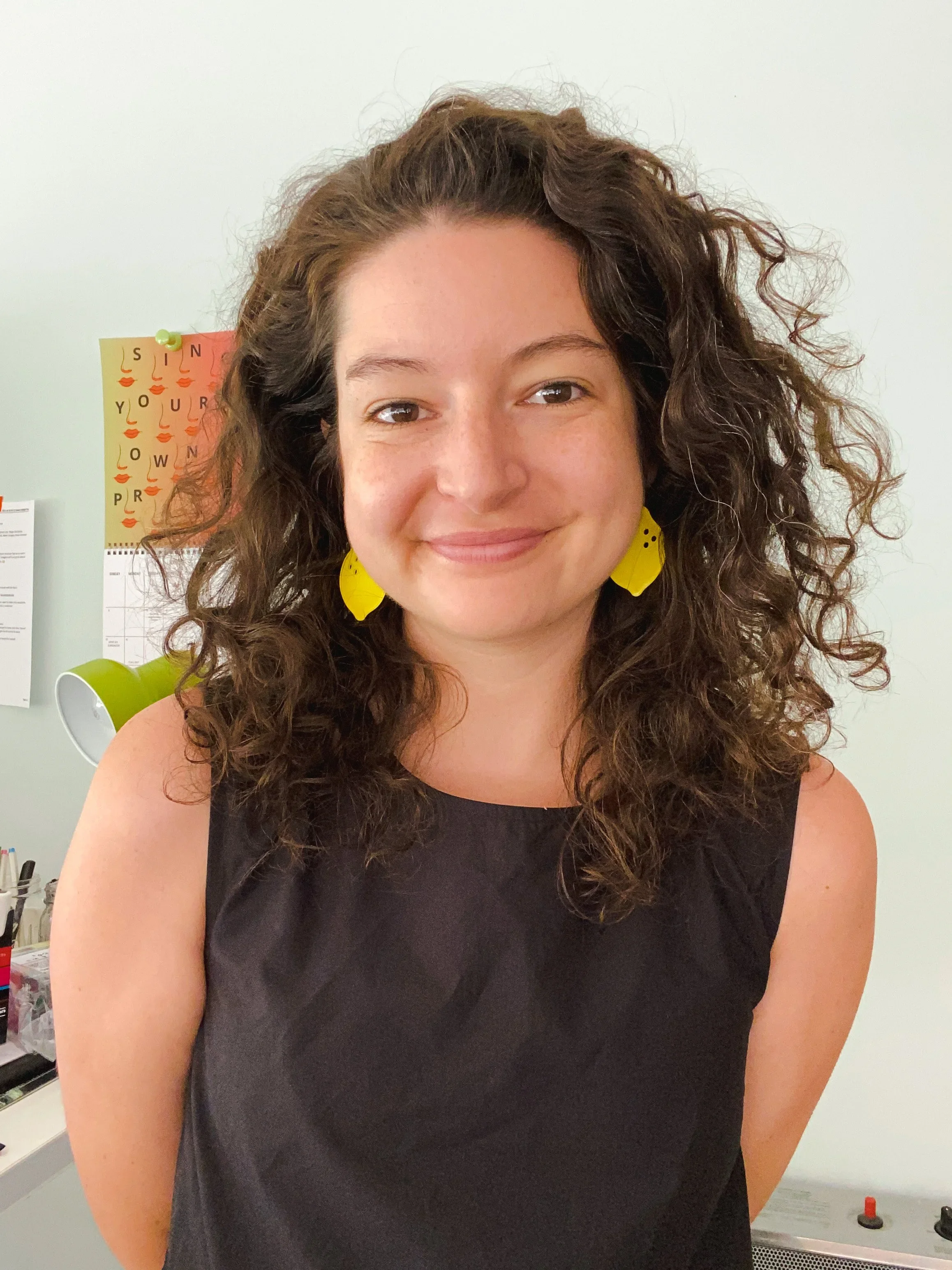 A woman with curly dark brown hair and yellow earrings smiling at the camera, standing indoors with a white wall and various papers in the background.