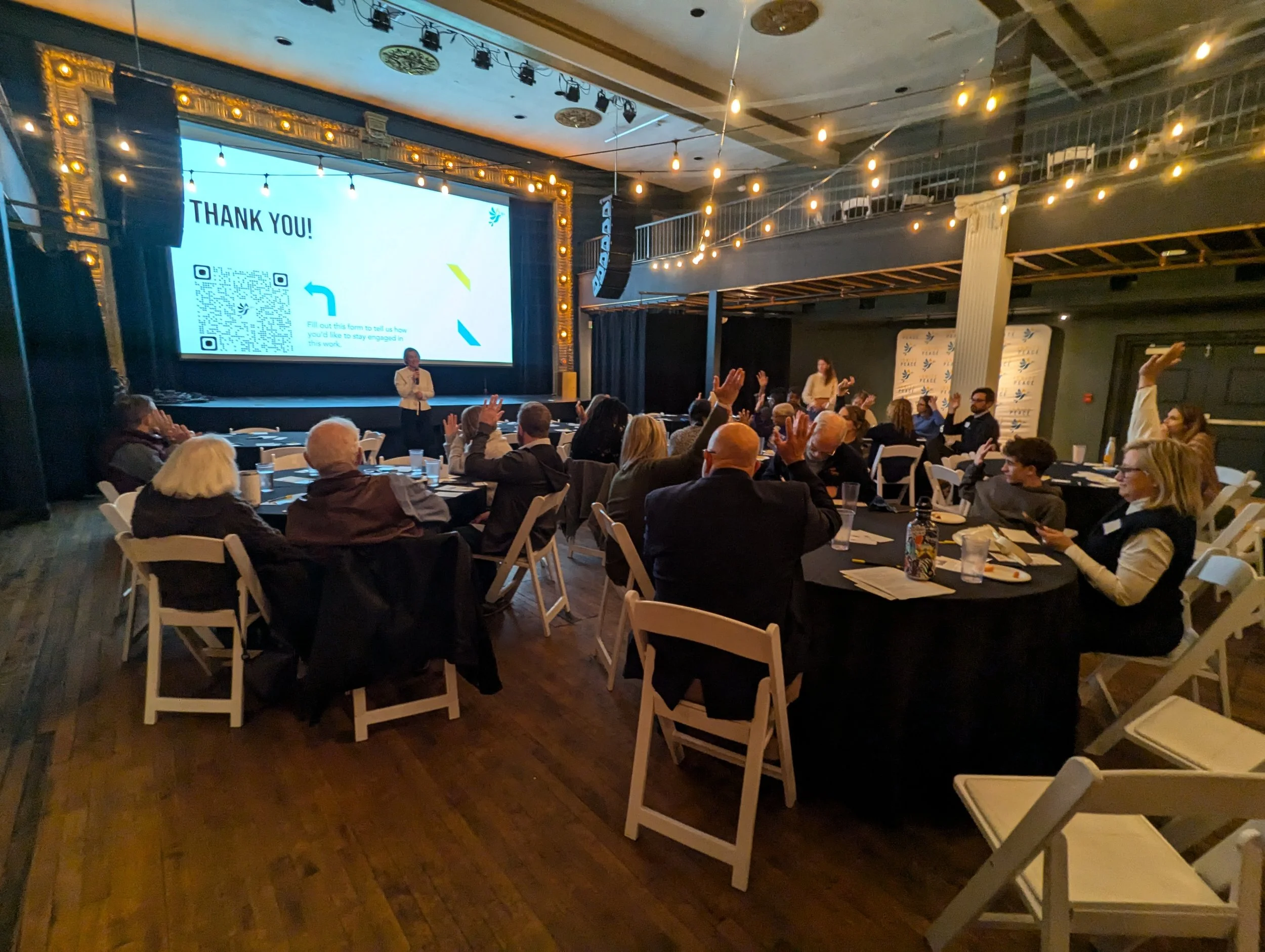 A group of people attending a conference or event in a dimly lit room with a large screen displaying a 'Thank You!' message. Several attendees have their hands raised while a presenter stands at the front near the screen.