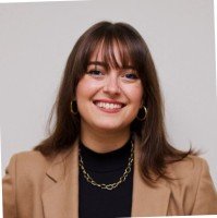 Portrait of a young woman with brown hair, wearing a tan blazer and black top, smiling, against a plain gray background.