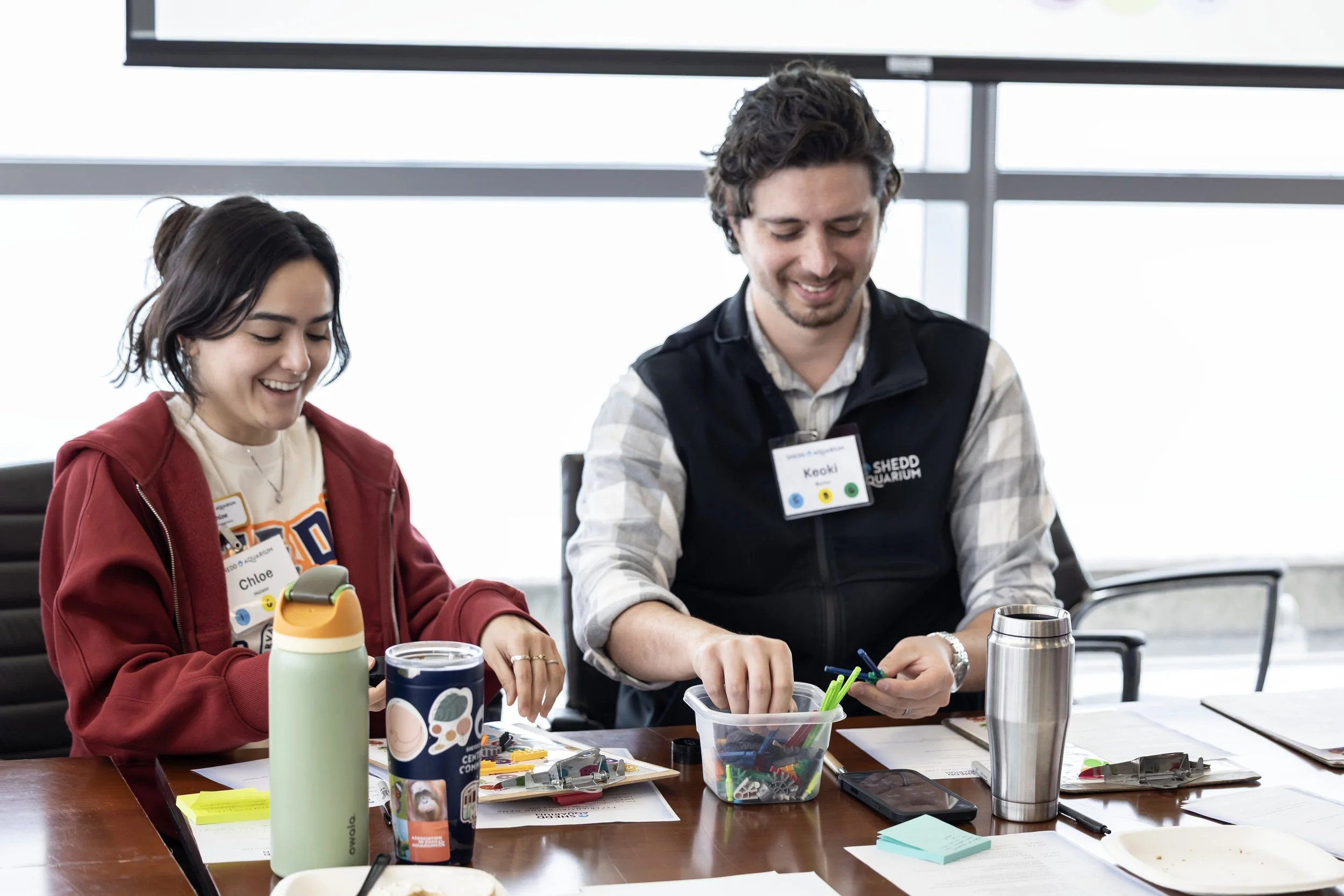 Two people sitting at a table working on creative projects, smiling, with various supplies, notebooks, and a stainless steel tumbler on the table.