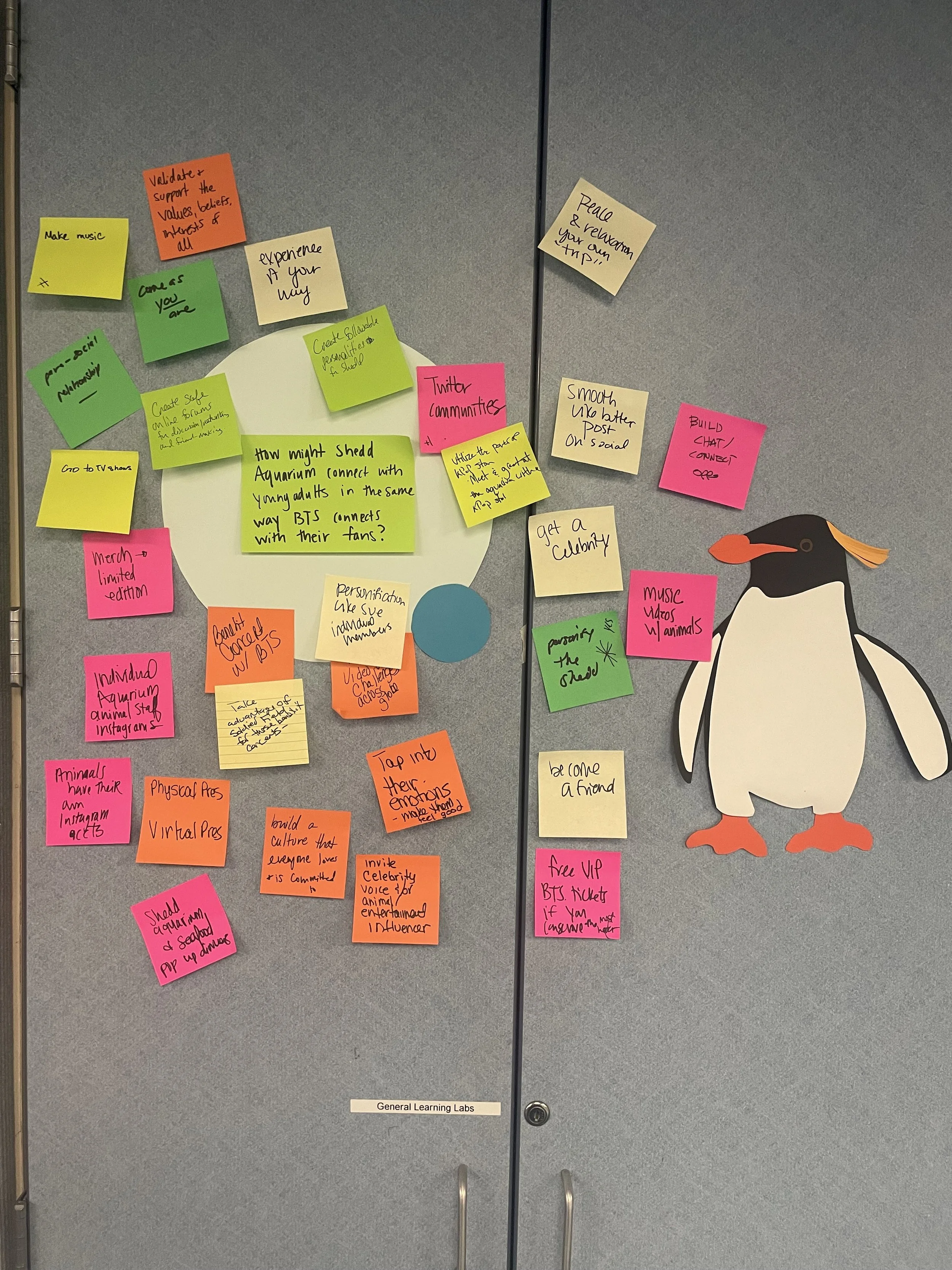Wall with colorful sticky notes and a paper penguin cutout. The notes contain ideas and questions related to community building, social media, and aquariums, such as 'Make music,' 'Twitter communities,' and 'Get a celebrity or influencer.'