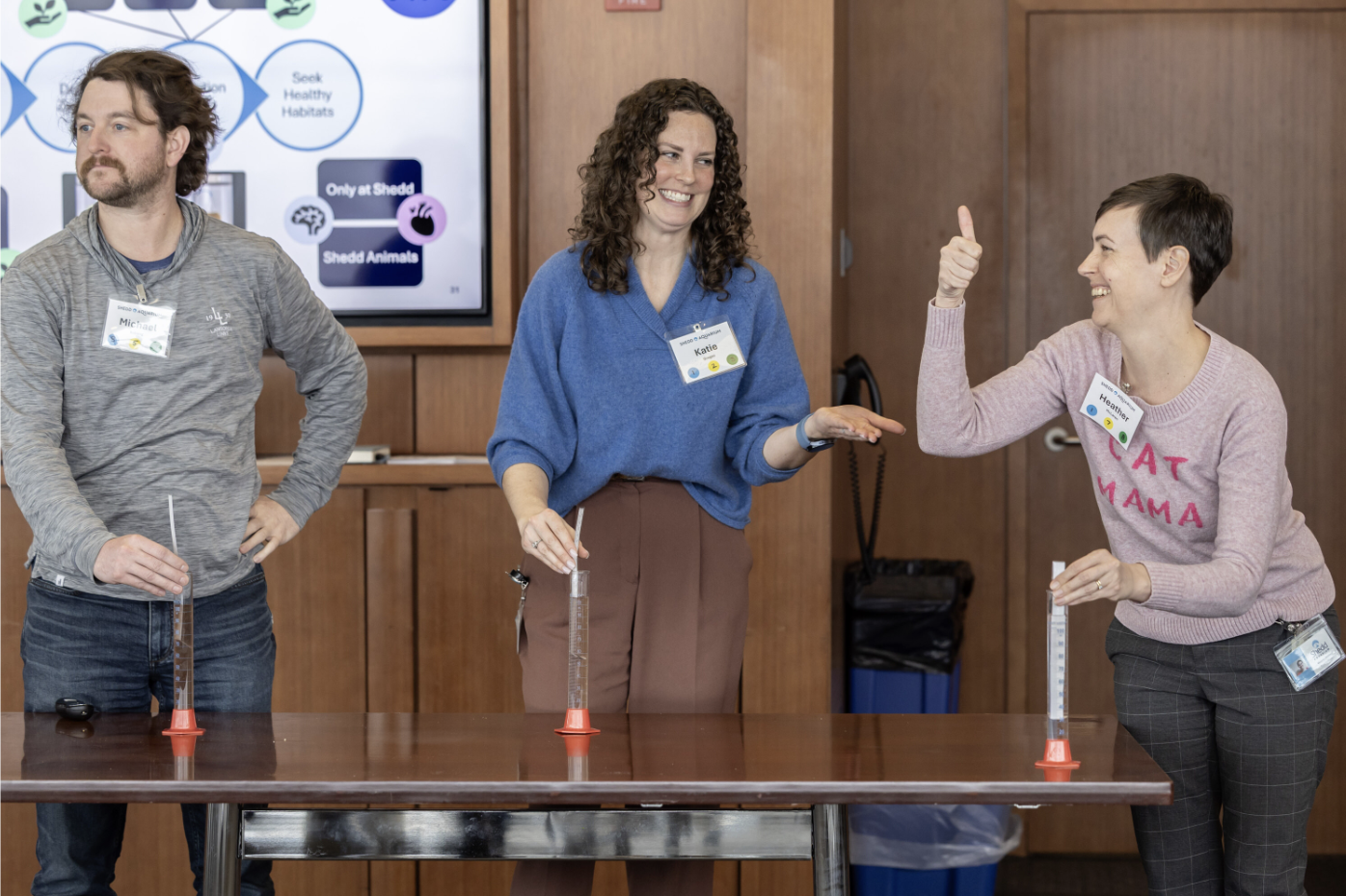 Three people stand around a table with measuring cylinders, indoors, smiling and engaging in conversation, with a wooden wall and a presentation screen in the background.