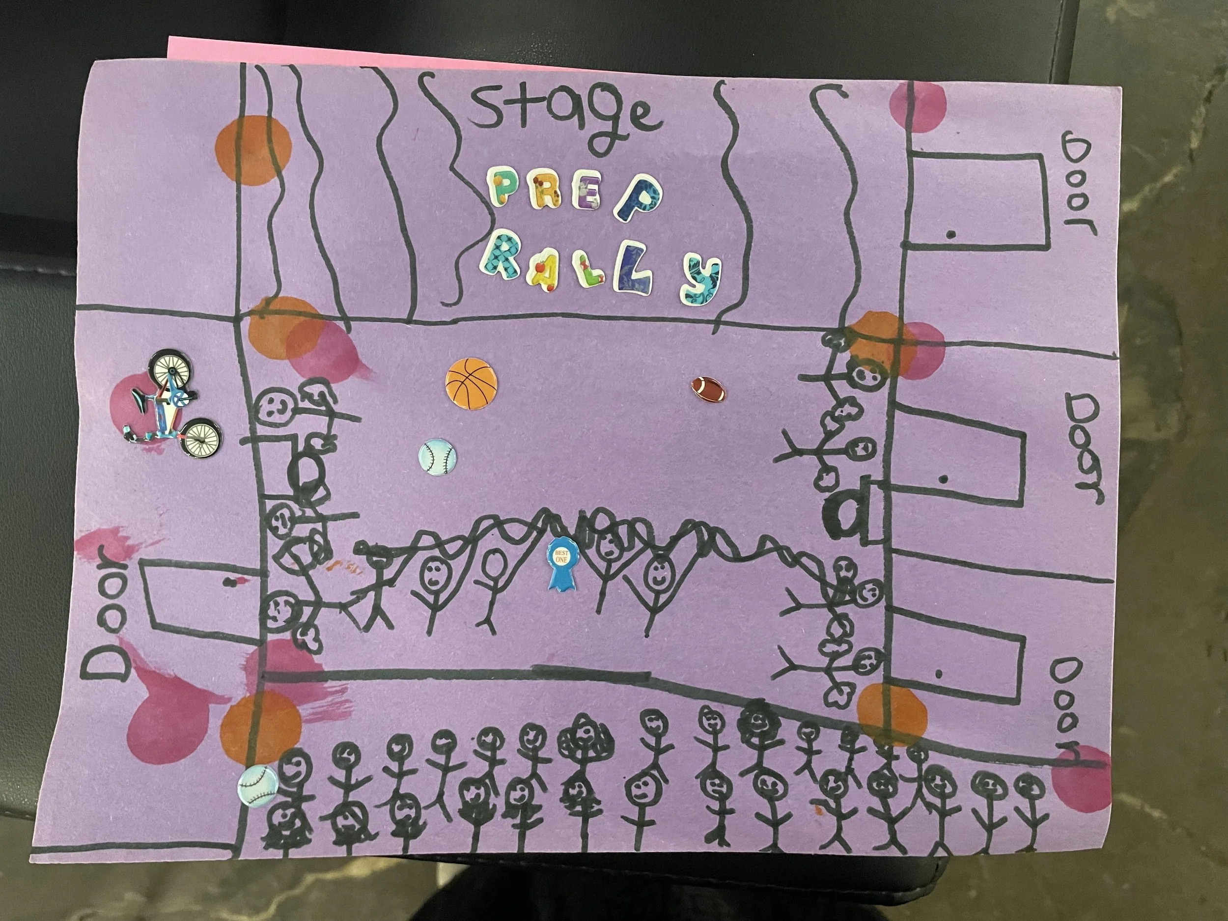 A hand-drawn poster on purple paper depicts a community event with a sports theme. It includes a central area labeled 'Stage' with colorful letter stickers spelling 'Prep Rally,' surrounded by drawings of people, sports balls, and sections labeled 'Door'. Stick figures represent participants, and various stickers of a bicycle, baseball, basketball, and football are attached to different parts of the poster.