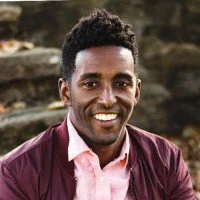 A smiling man with dark curly hair, wearing a pink shirt and maroon jacket outdoors with a rocky background.