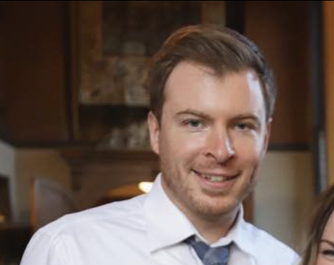 A young man with light brown hair and a beard, smiling, wearing a white shirt and a tie, in a cozy indoor setting.