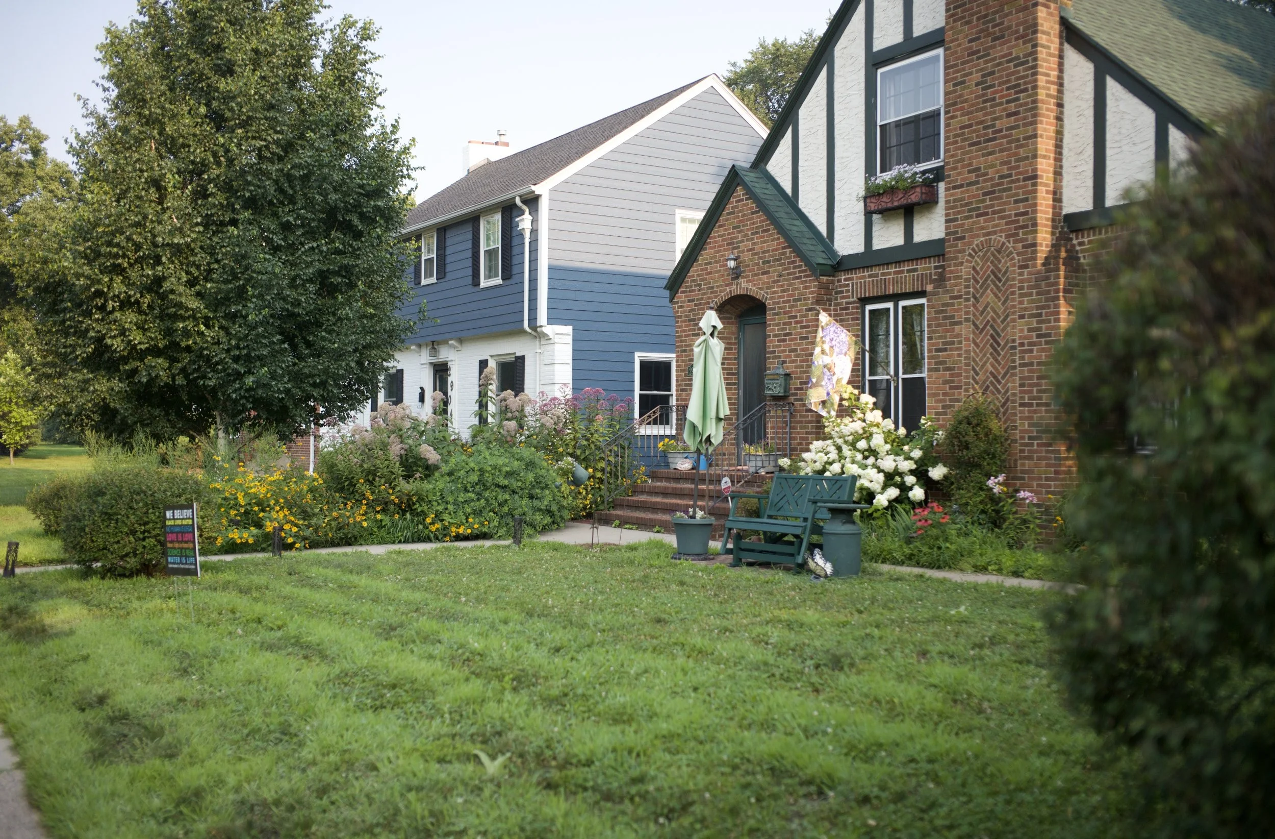 Front yard of a suburban house with green grass, colorful flowers, garden bushes, and trees, with neighboring homes visible in the background.