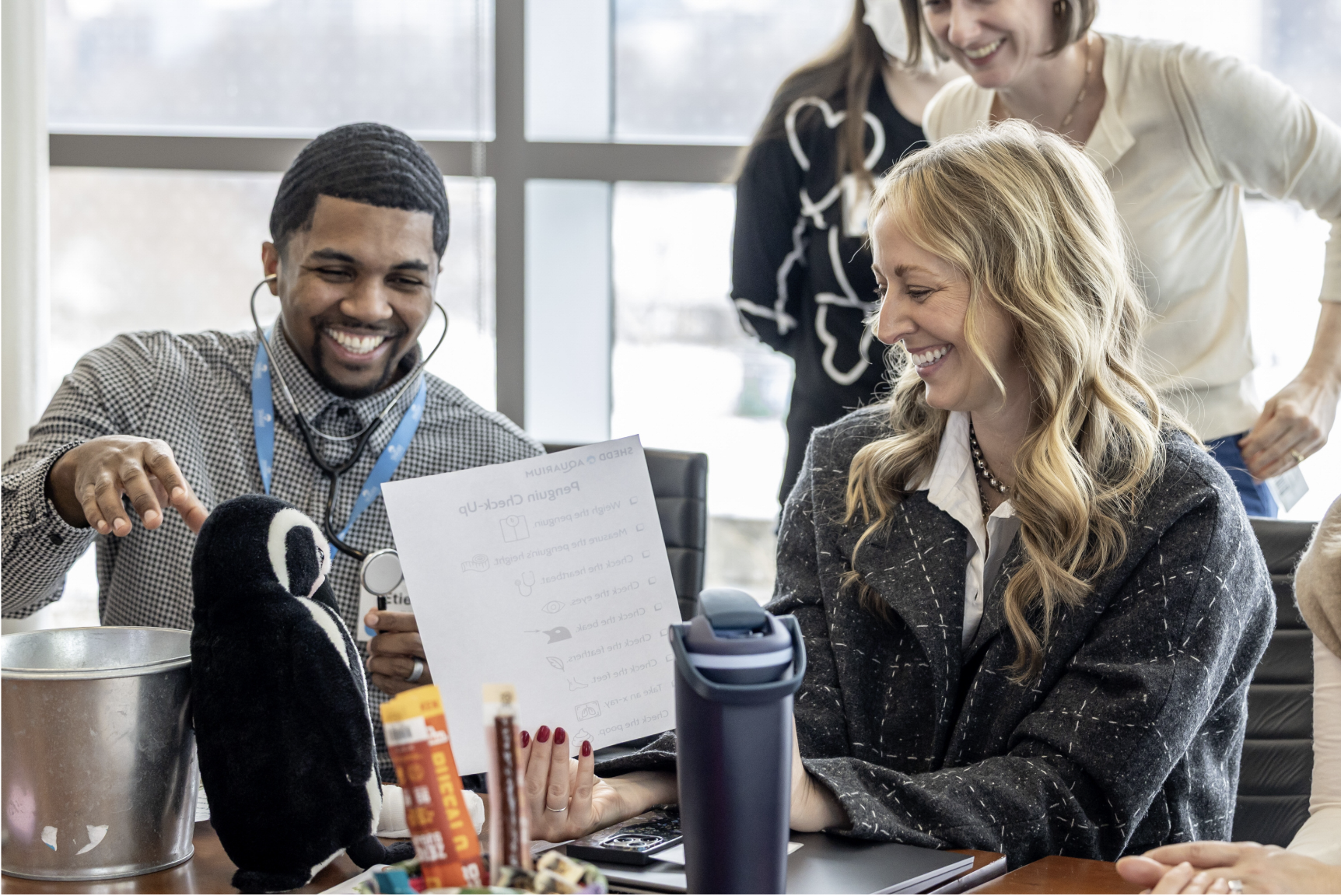 Group of smiling people participating in a workshop, with a woman petting a small black and white plush penguin in an office setting.