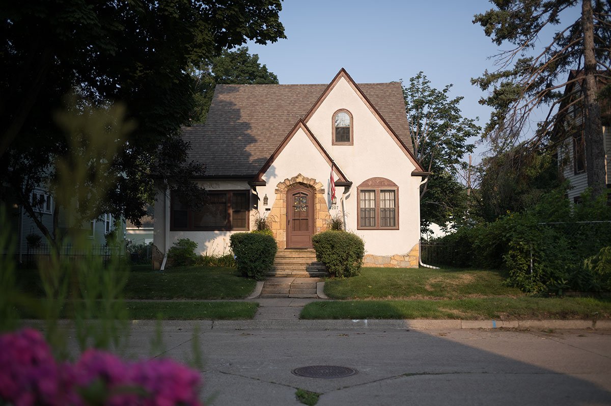 A house with a steep roof, stone steps leading to a wooden front door, surrounded by greenery, under a clear sky during sunset.