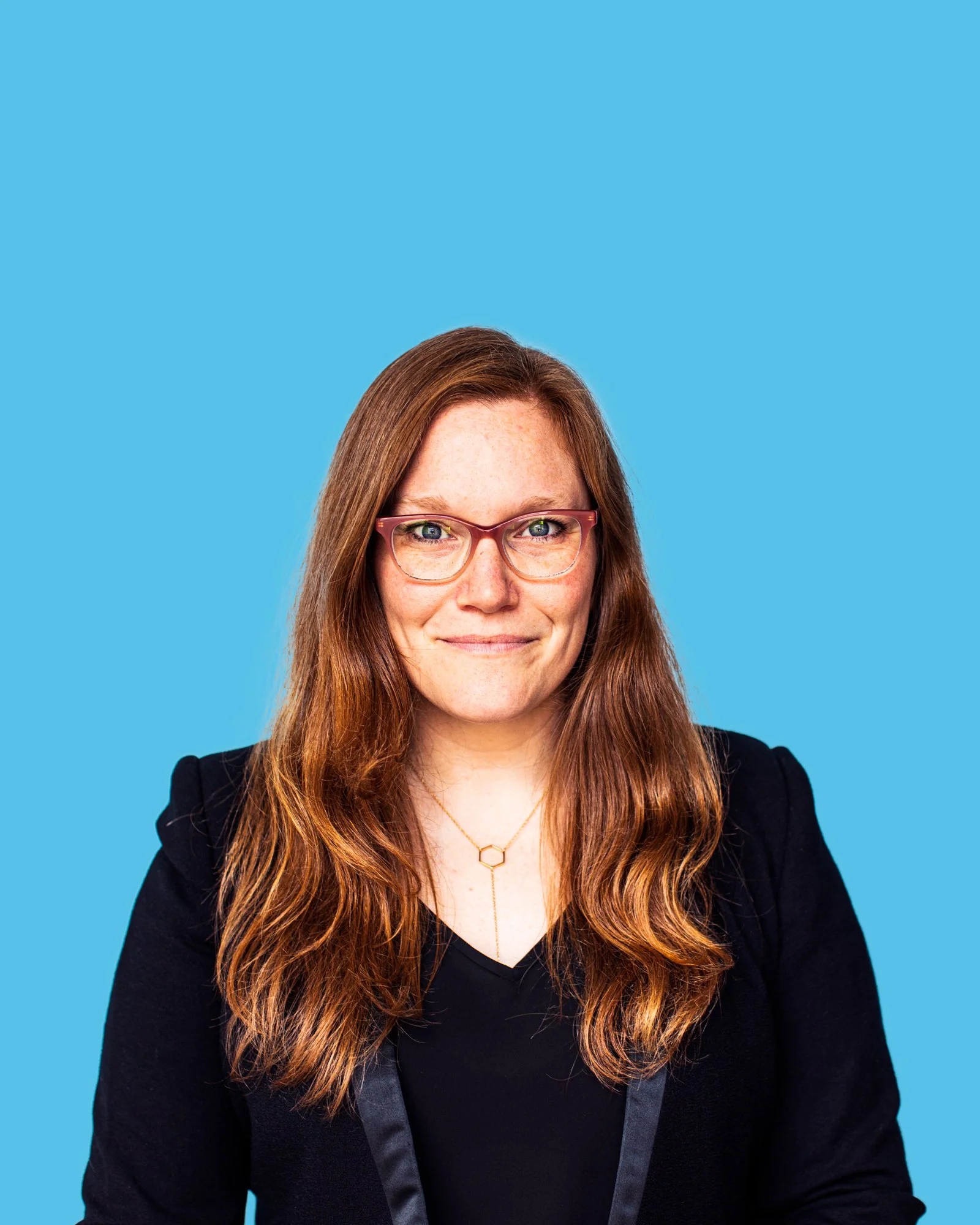 Woman with long reddish-brown hair, glasses, wearing a black blazer, smiling, against a blue background.