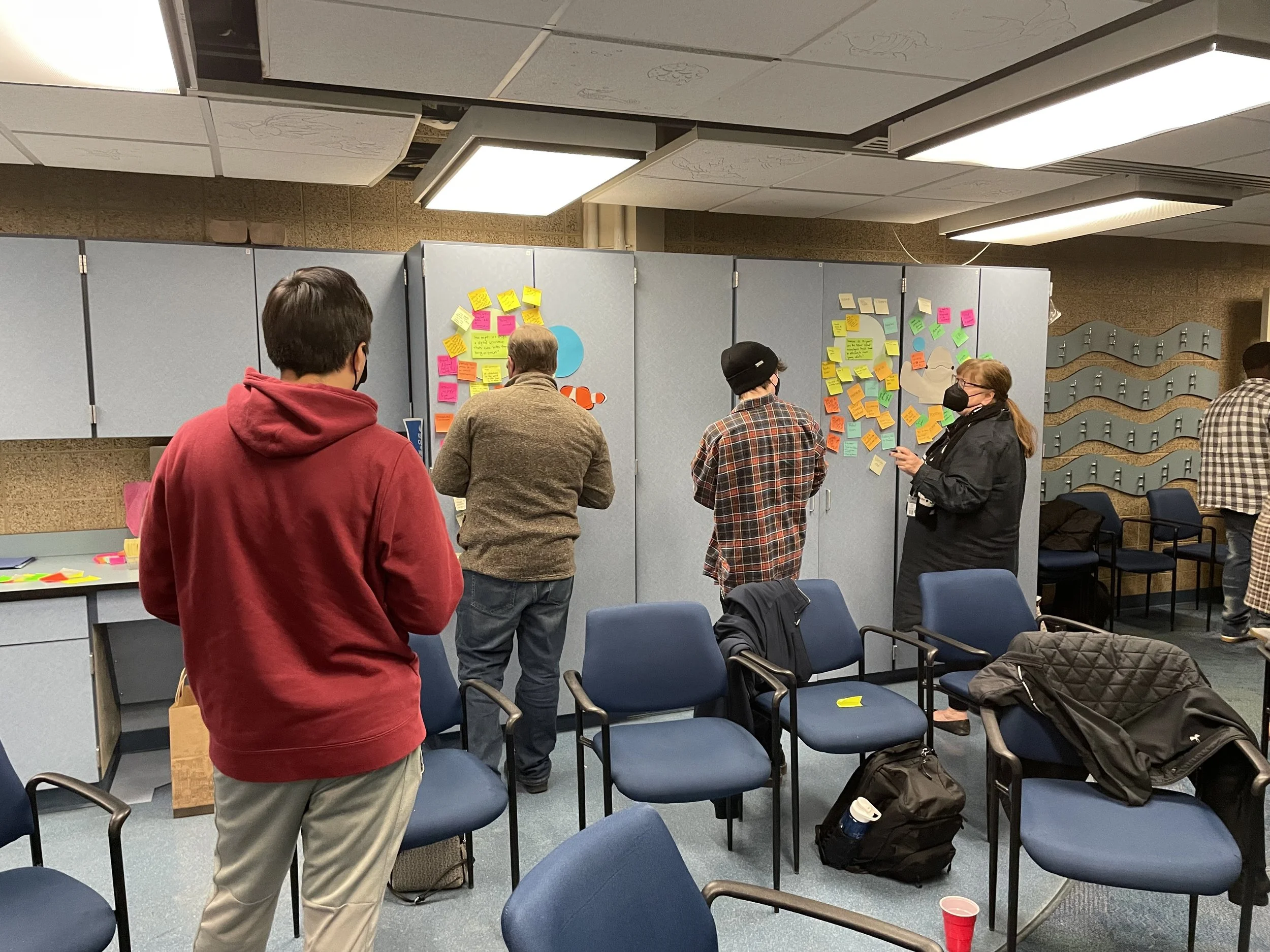 People standing and sitting in a room with colorful sticky notes on the walls, engaging in a group activity or discussion.