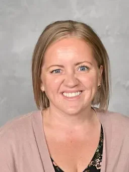A woman with short blonde hair smiling at the camera, wearing a light pink cardigan over a floral top, standing against a gray background.