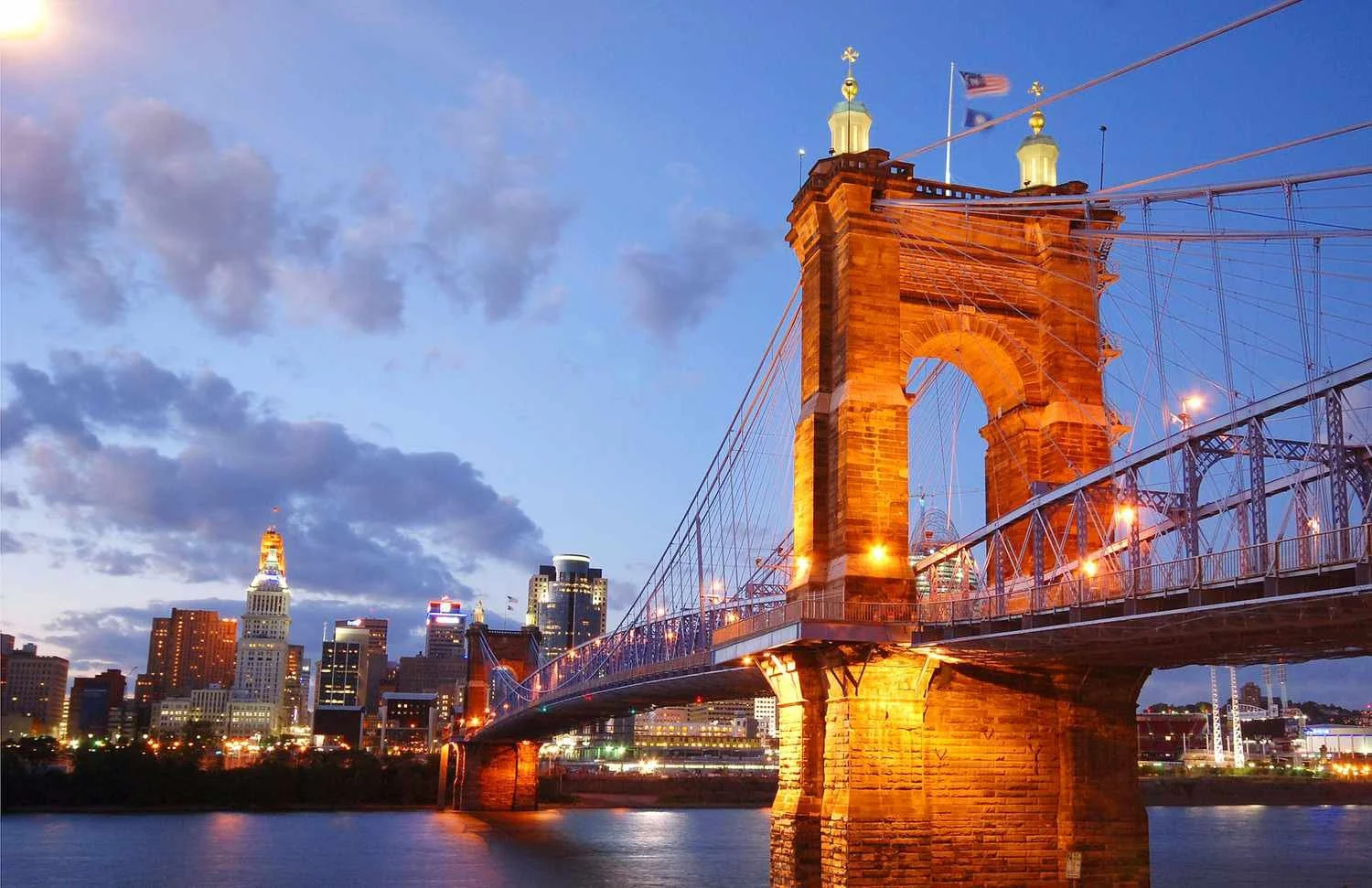 The illuminated John A. Roebling Suspension Bridge over the Ohio River with Cincinnati skyline in the background at dusk.
