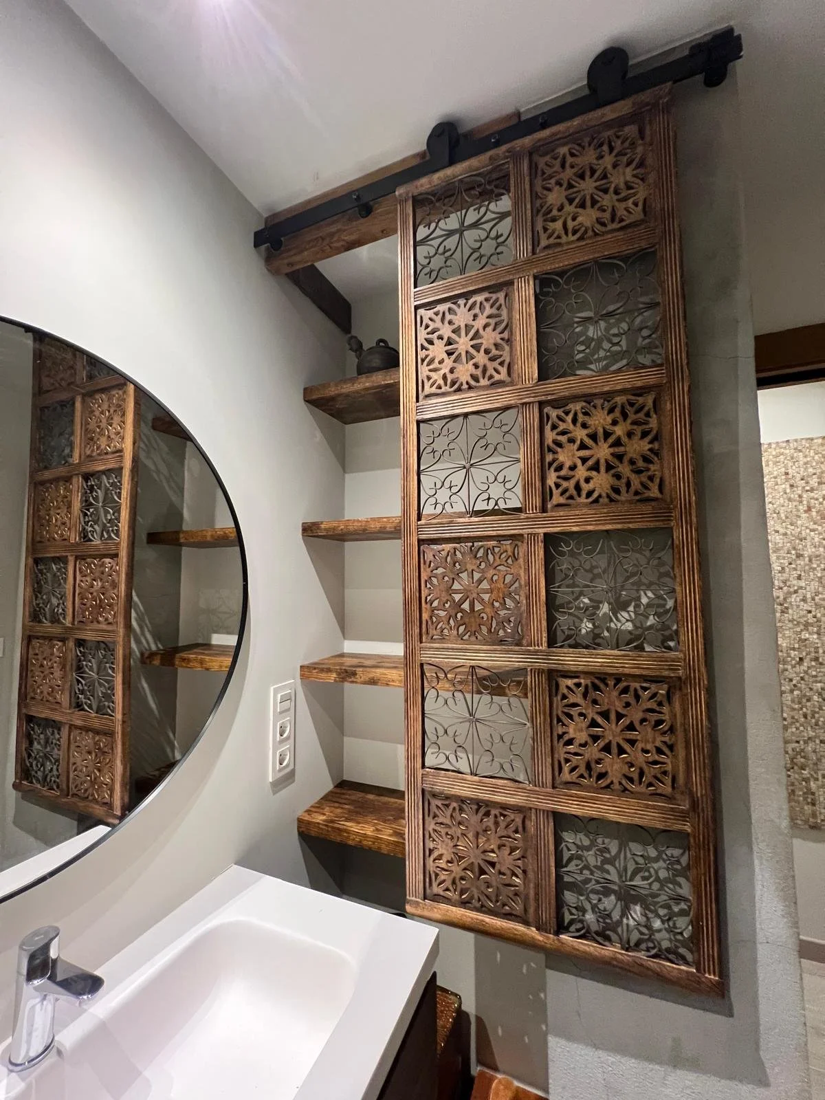 A bathroom with a white sink, a circular mirror, and decorative wooden shelves with intricate carved panels, mounted on a gray wall.