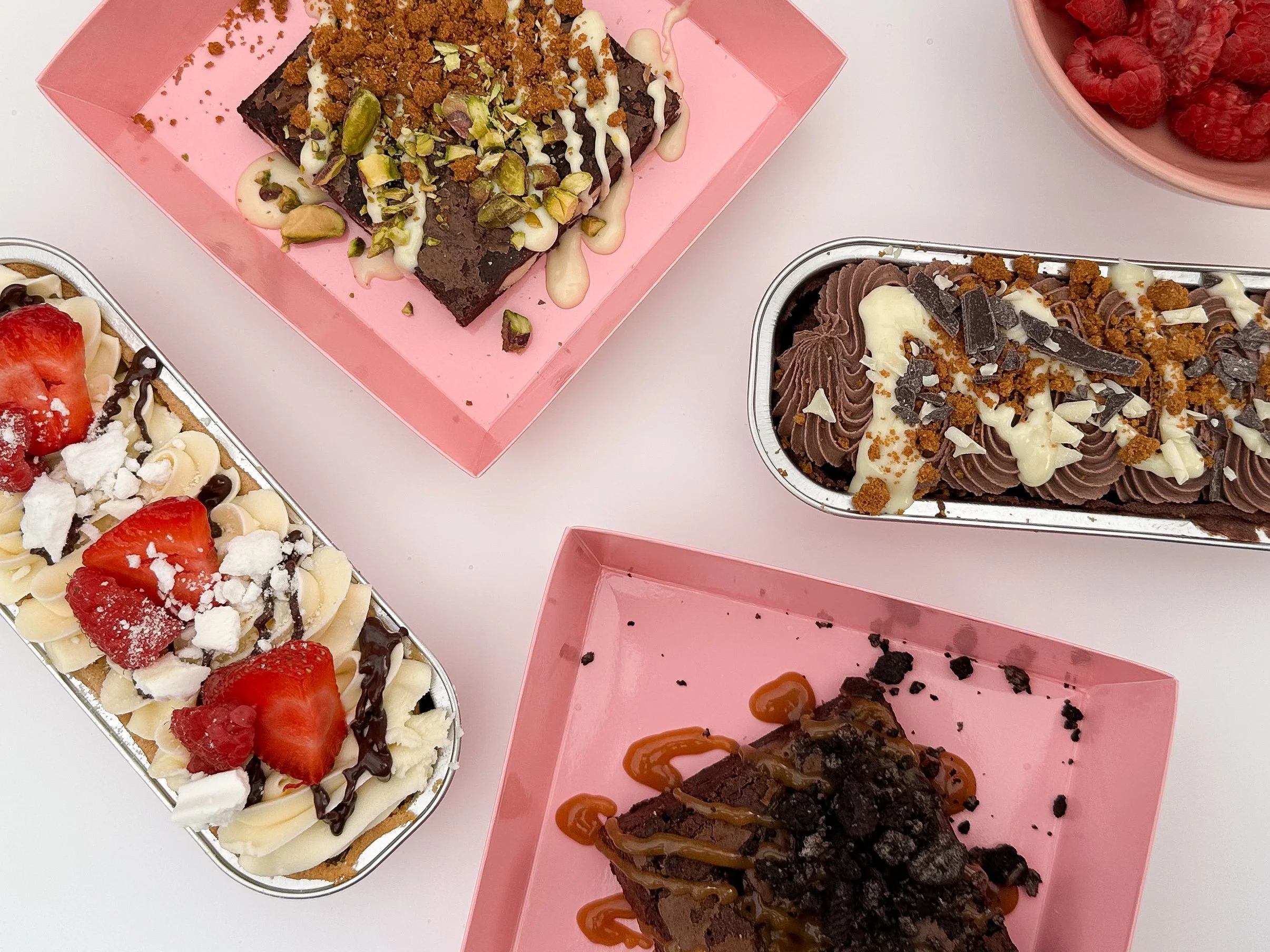Assorted decorated brownies with various toppings such as strawberries, raspberries, chocolate chunks, pistachios, and caramel drizzle, served on pink platters.
