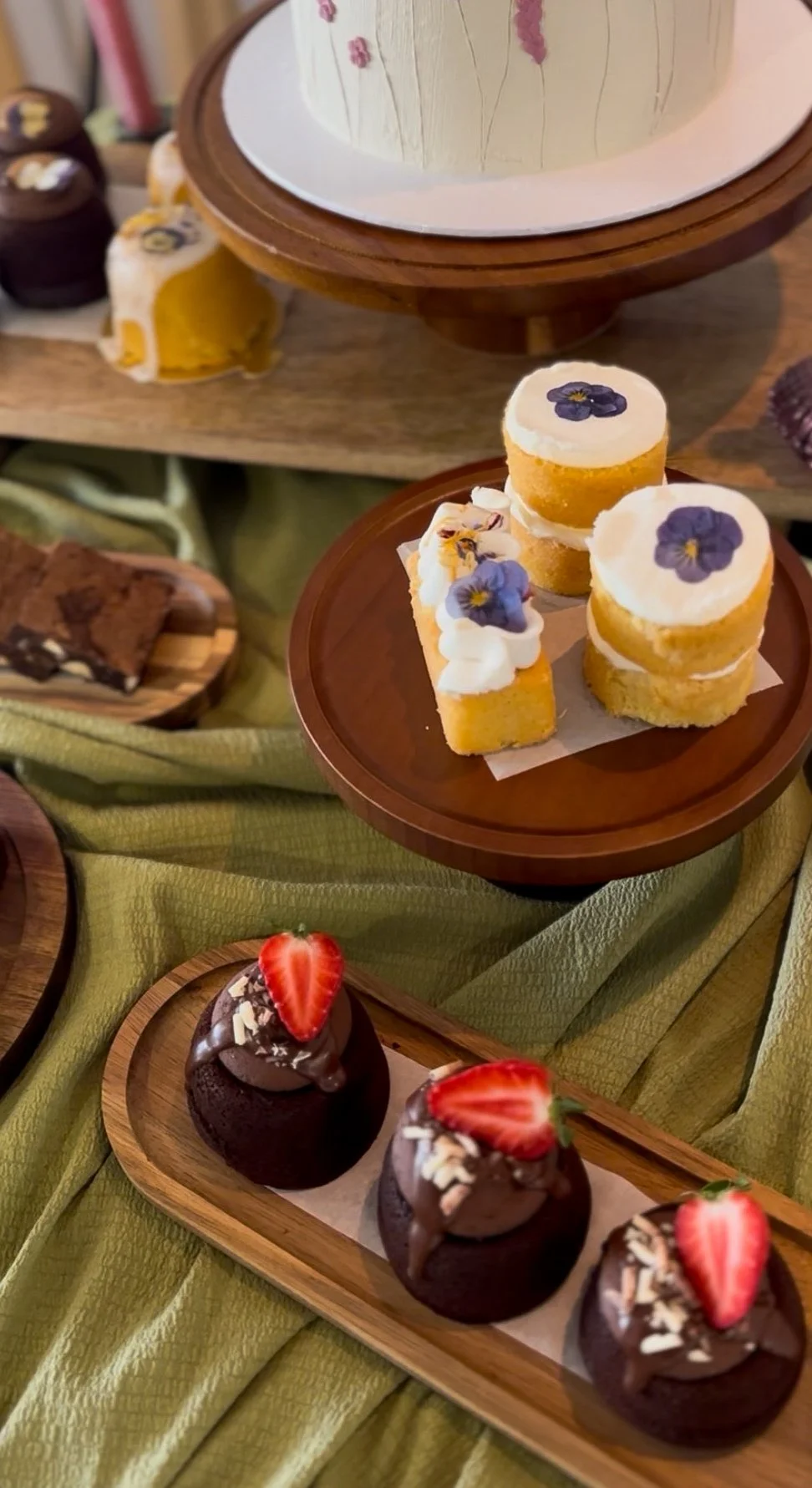 Assorted cakes and desserts displayed on wooden and ceramic plates, including mini layered cakes with cream and flower decorations, chocolate cupcakes topped with strawberries and white chocolate shavings, all arranged on a green fabric surface.