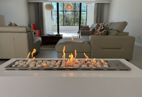Modern living room with beige sofas, colorful pillows, large windows with curtains, and a floating gas fireplace with flames and decorative rocks.