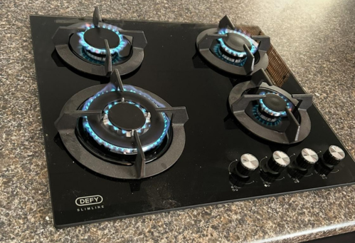 A black four-burner gas stove with blue flames on all burners, placed on a dark countertop.