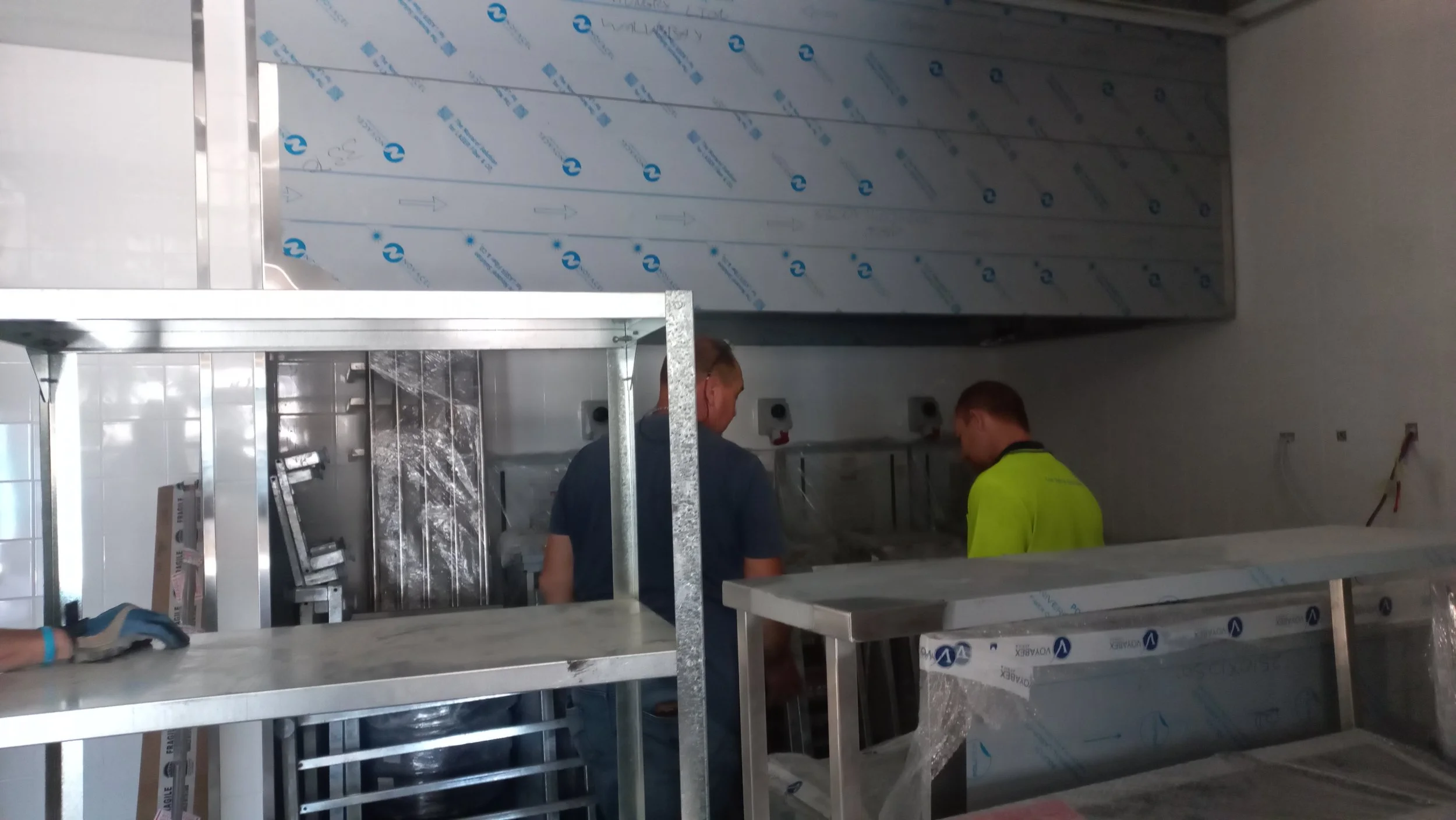 Two workers in a kitchen, one in a dark shirt and the other in a bright yellow shirt, working on installing or assembling kitchen equipment, with stainless steel counters and an appliance still wrapped in protective plastic.