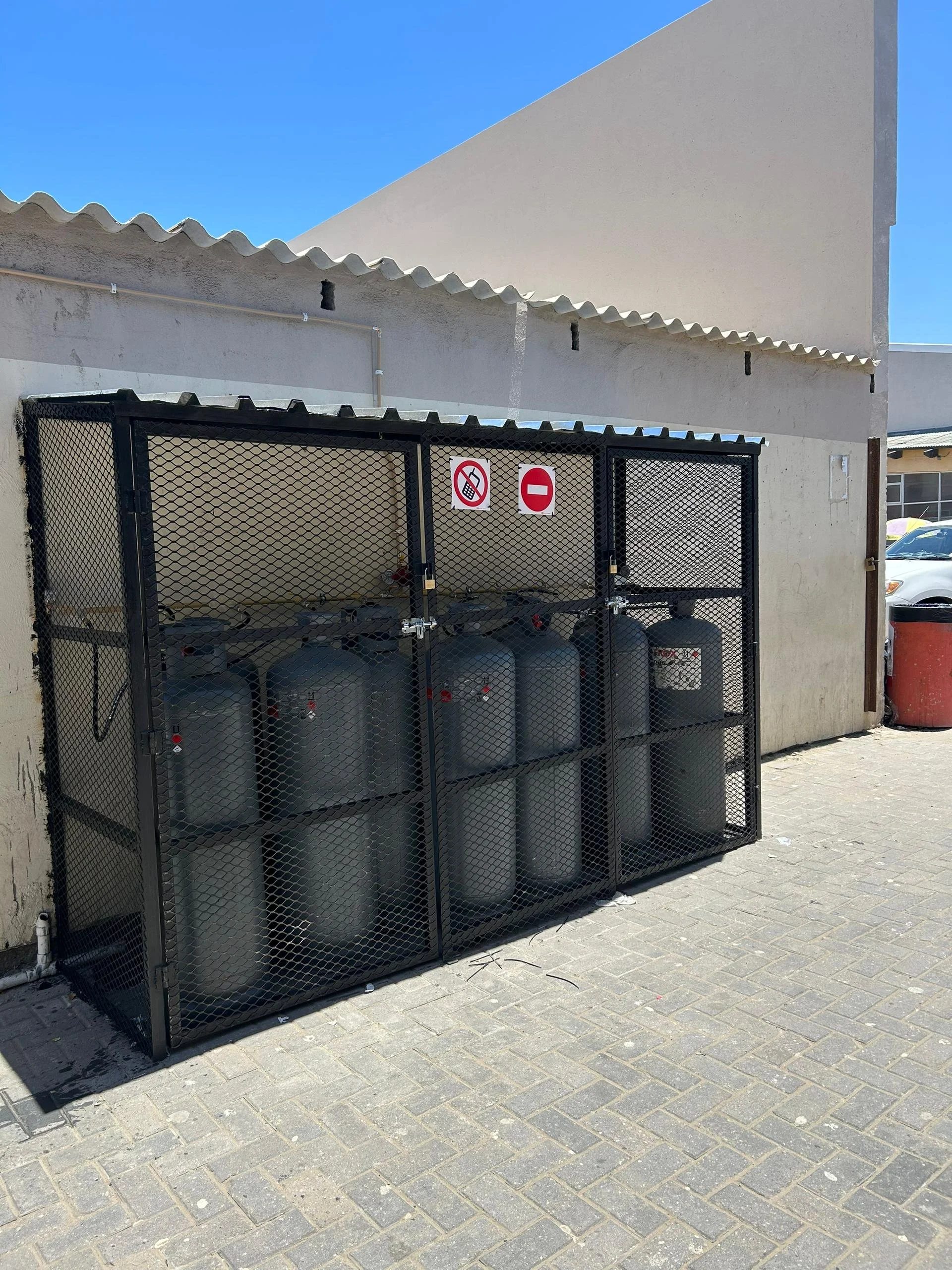 A black metal cage containing multiple grey gas cylinders, located outdoors on a paved area against a beige wall. There are signs on the cage indicating 'no smoking' and 'no entry'.