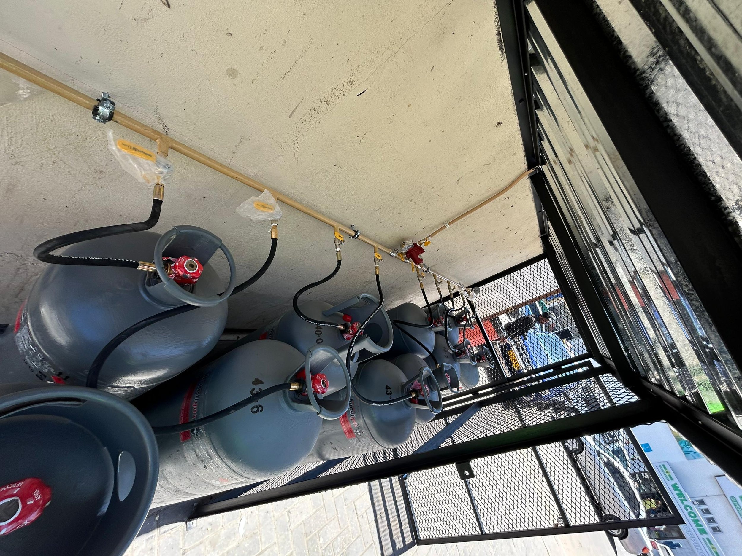 LP Gas cylinders stored under a metal roof, connected to a gas line, with a view of the parking area in the background.