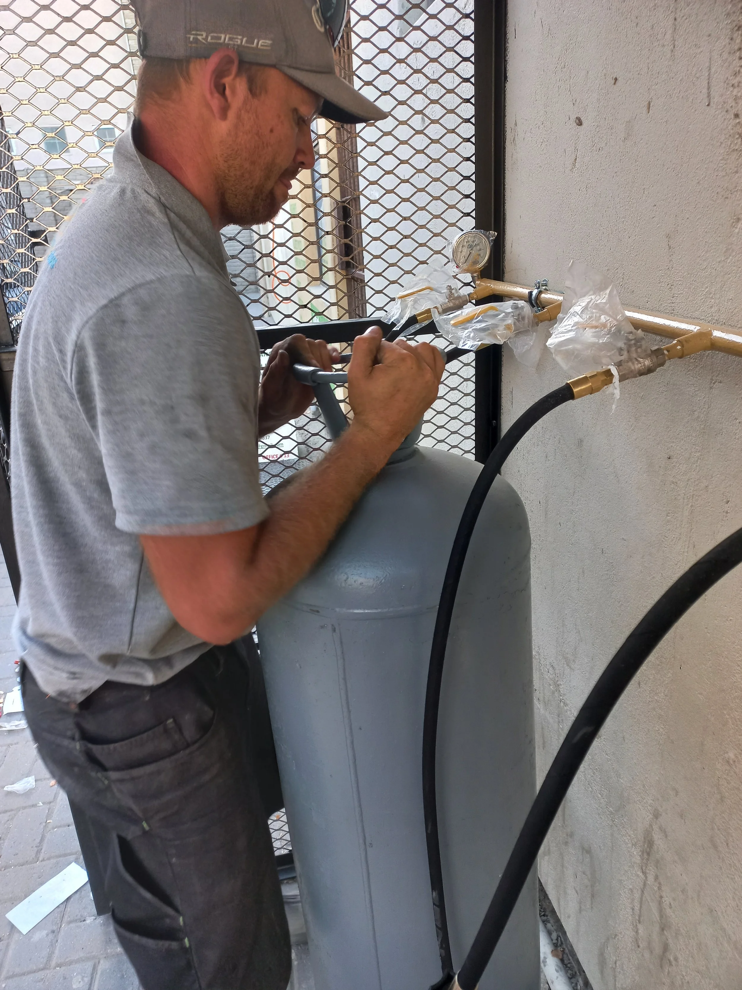 A technician is working on a gas cylinder connected to a set of pipes and gauges, possibly performing maintenance or repair.