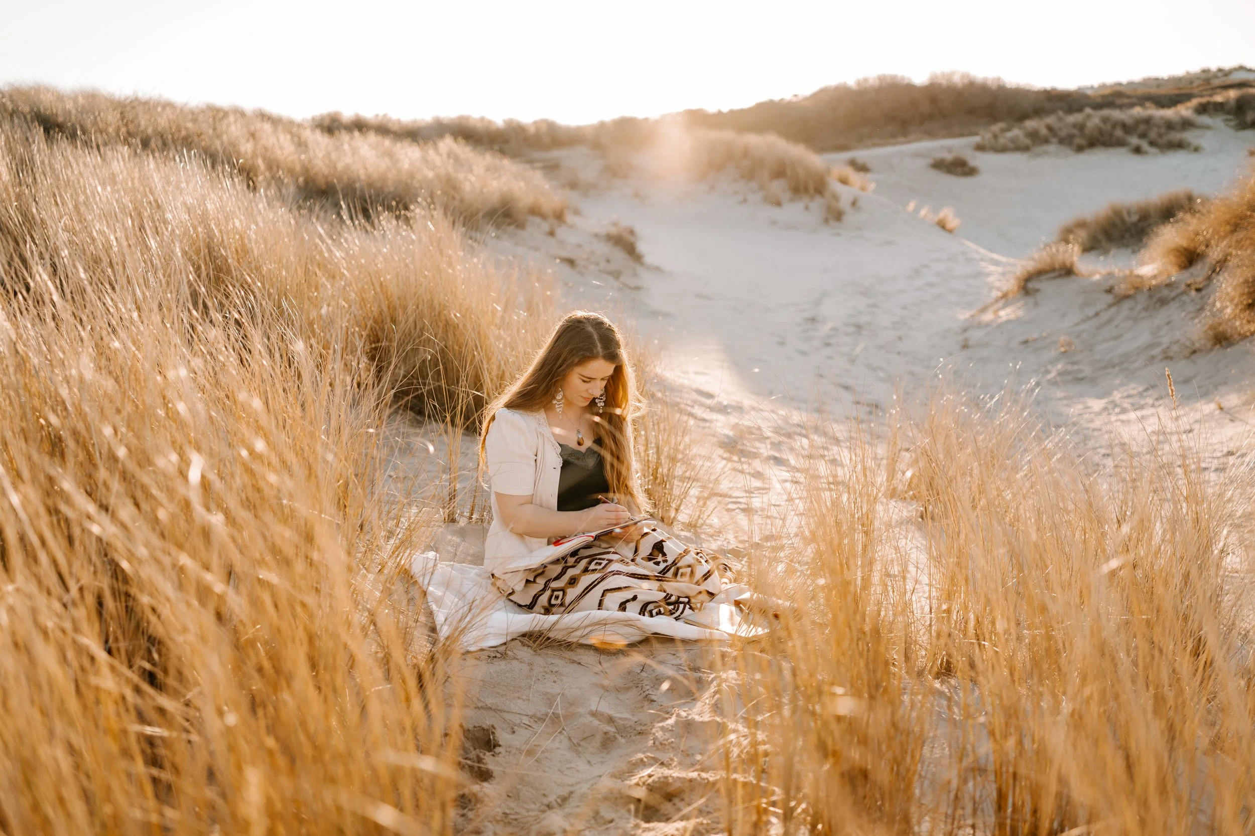 A woman sitting on the sand in a sandy area with tall grass, writing or drawing in a notebook, with the sunlight in the background.
