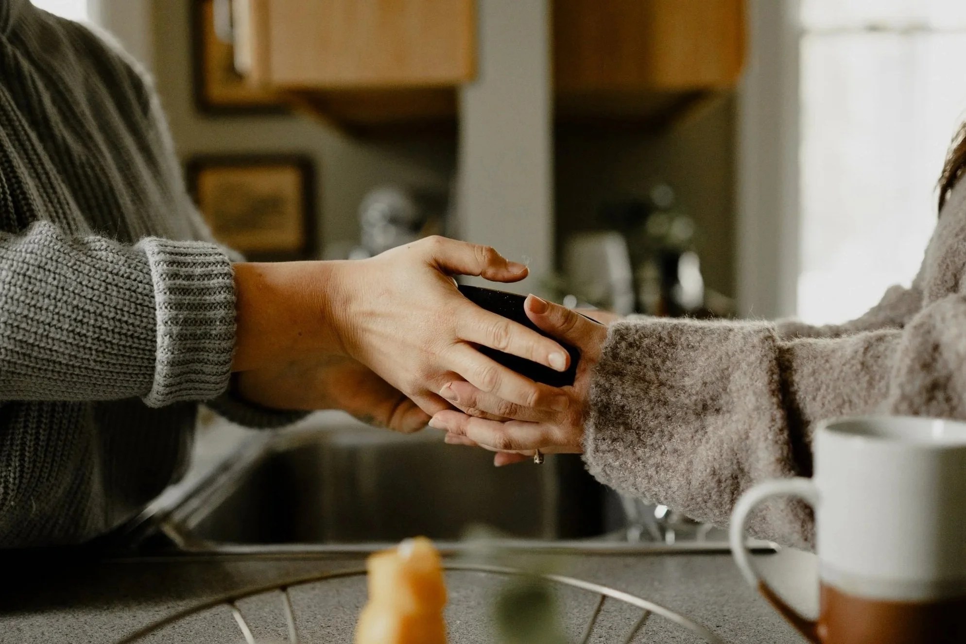 Two people holding hands, one person wearing a gray sweater and the other in a fuzzy beige sweater, in a kitchen setting.