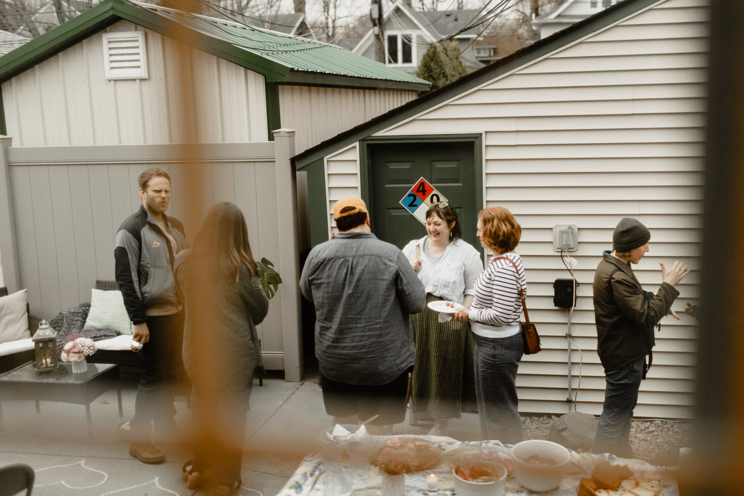 Group of people gathered outside in a backyard, some smiling and talking near a house with white siding, with food and drinks on a table in the foreground.