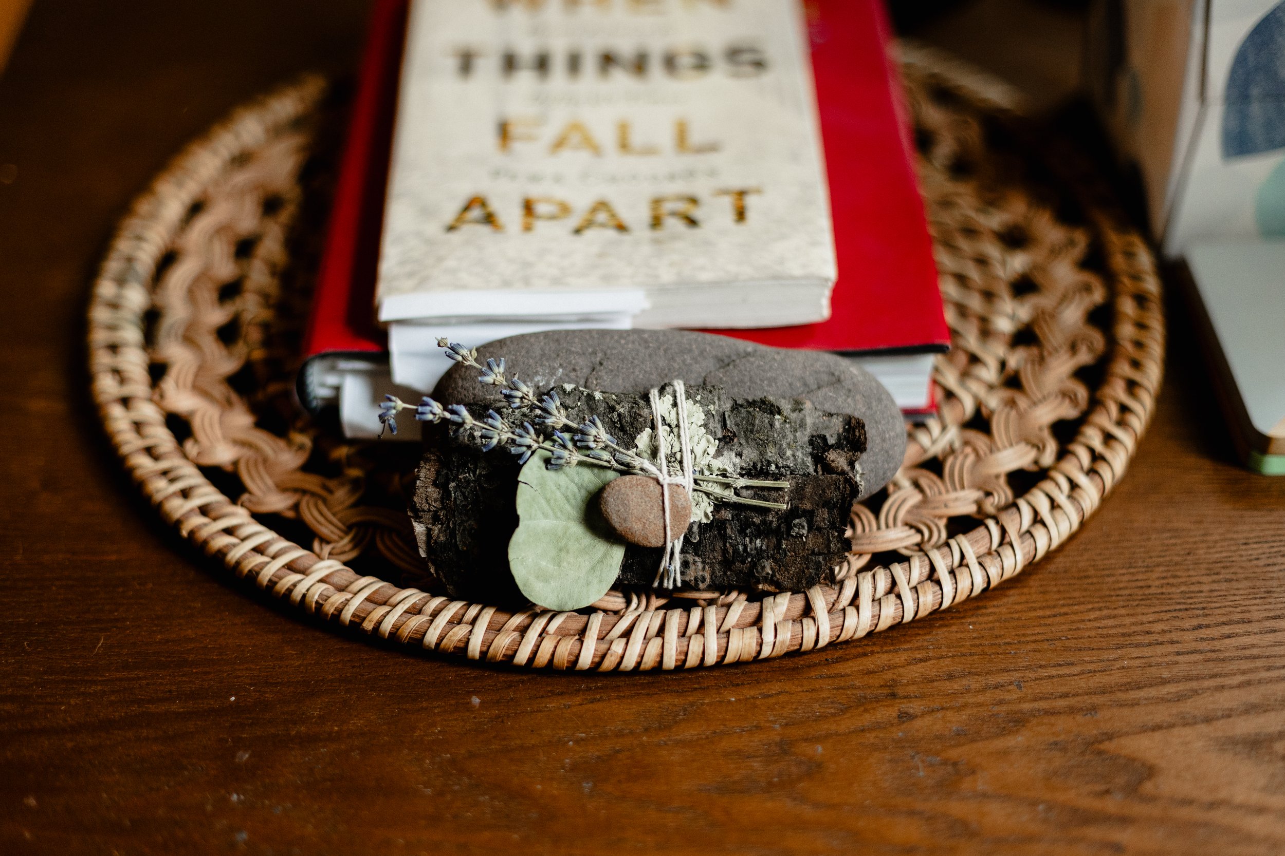 A wicker tray on a dark wooden surface containing a rock, a sprig of dried lavender, a green leaf, and small stones, with a blurred book in the background.