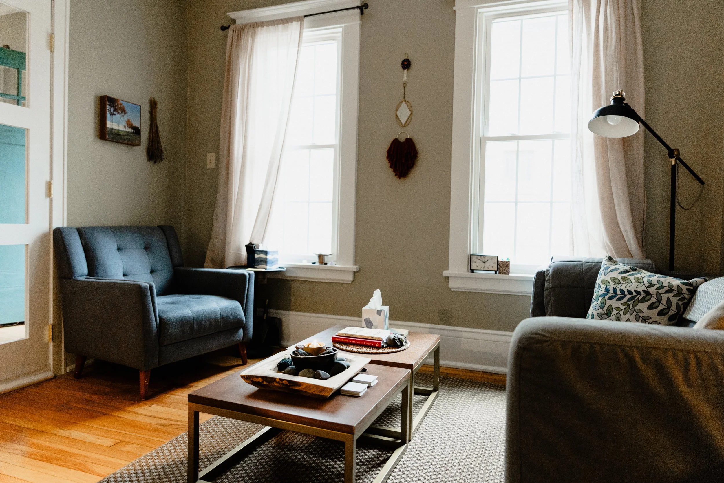 A cozy living room with two large windows with cream-colored curtains, a framed picture on the wall, a black armchair, a lamp with a black shade, a gray sofa with patterned pillows, a wooden coffee table with books and decorative items, and a stone t