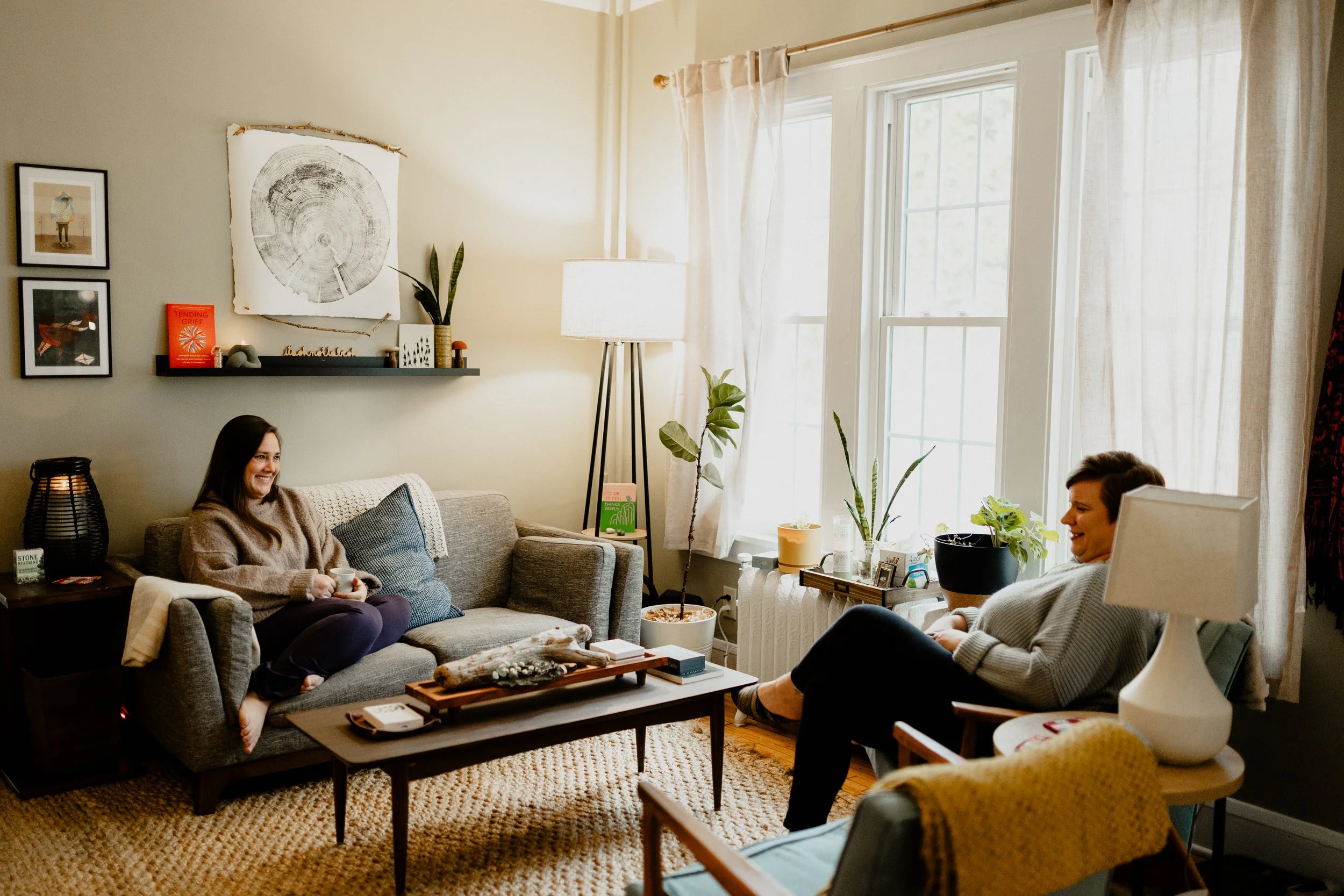 Two women sitting in a cozy living room, smiling and chatting, with plants and artwork on the wall, and sunlight coming through large windows.