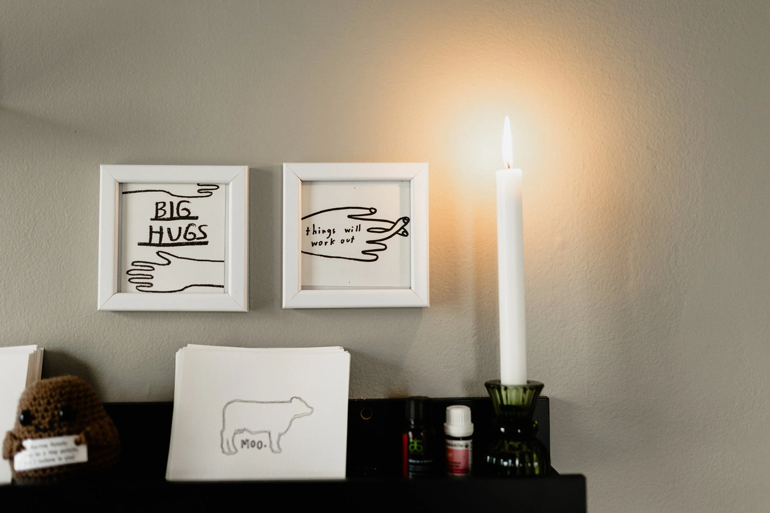 Decorative wall with two framed art pieces featuring hands and motivational texts, a lit white candle, and small essential oil bottles on a black surface, with a crocheted stuffed animal in the foreground.