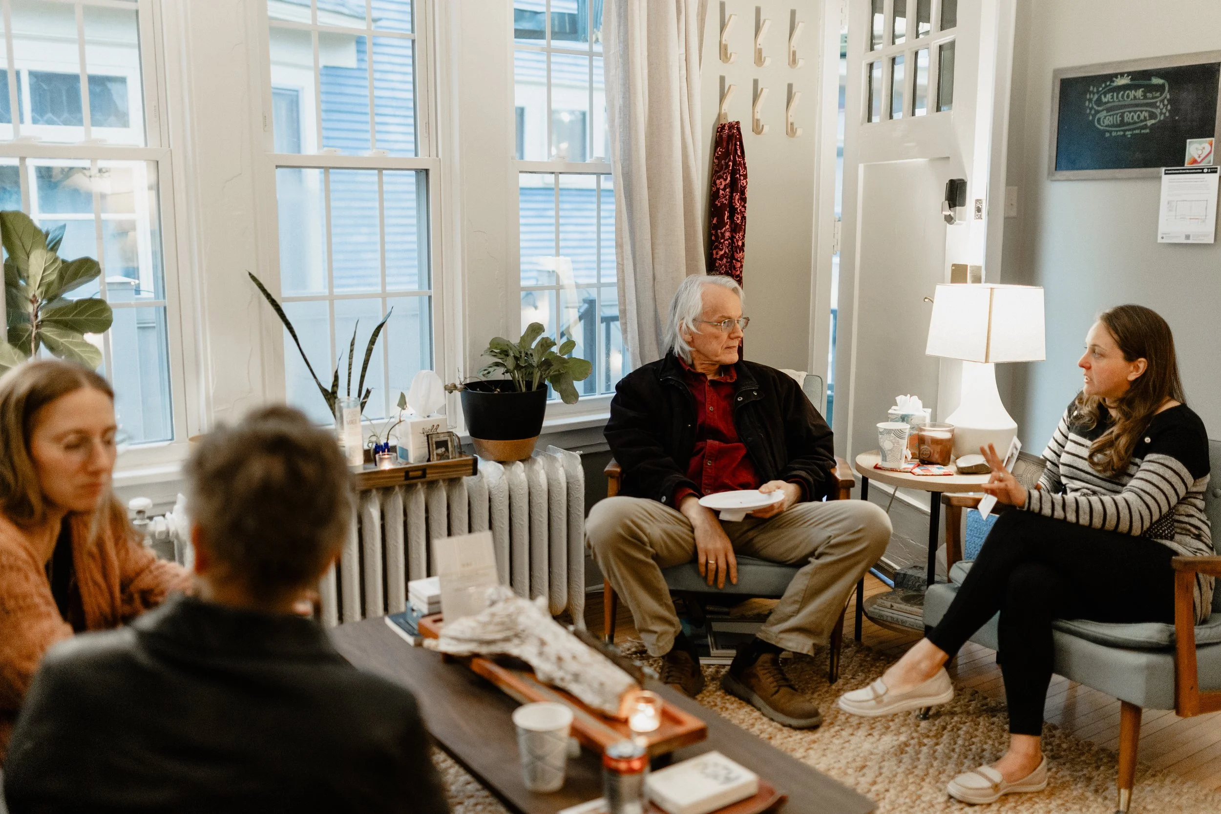 People sitting in a cozy room with large windows, plants, and a lamp, engaging in conversation.