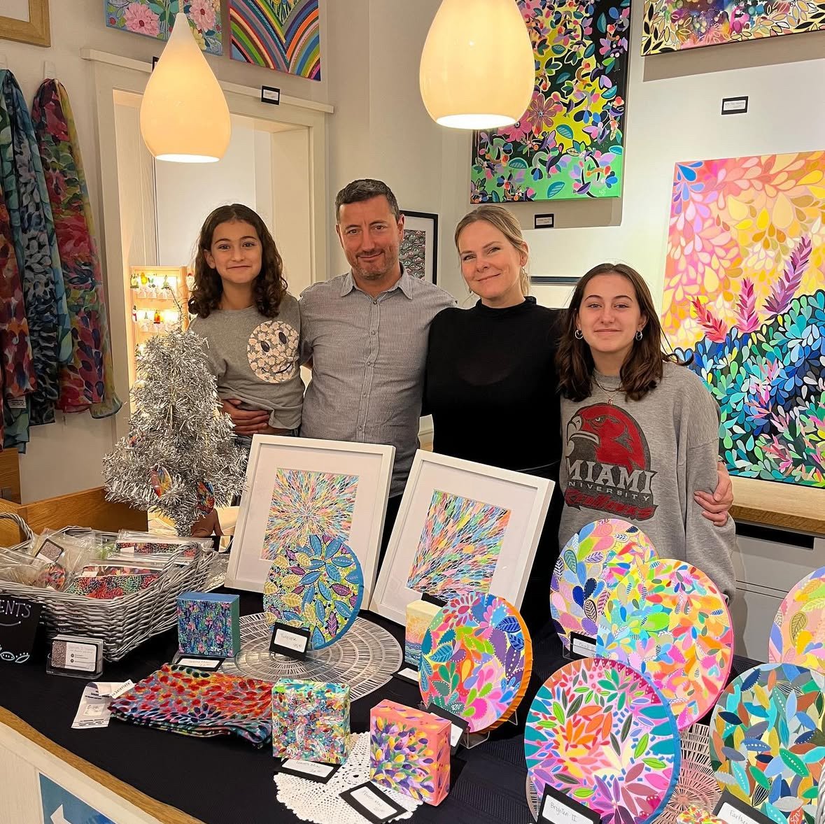 Artist Sarah J. Vanni stands with her husband and two daughters, surrounded by a display of her vibrant and colorful works of art.