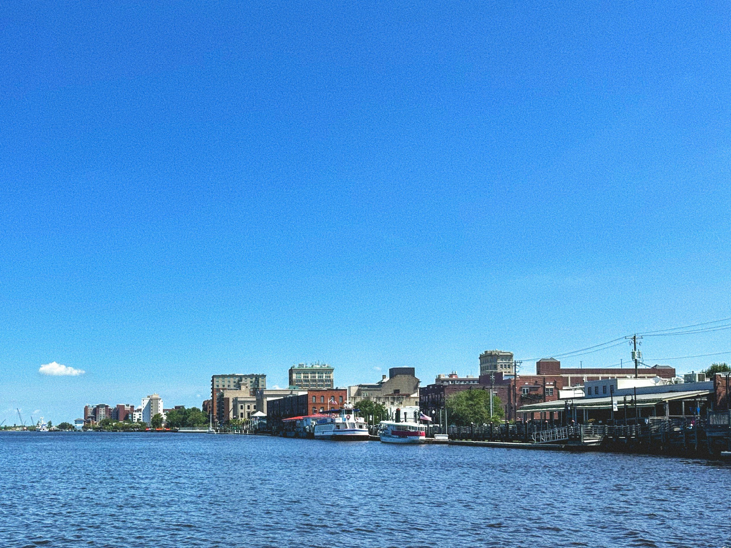 View of a city waterfront with boats docked along the pier and a row of buildings against a bright blue sky.