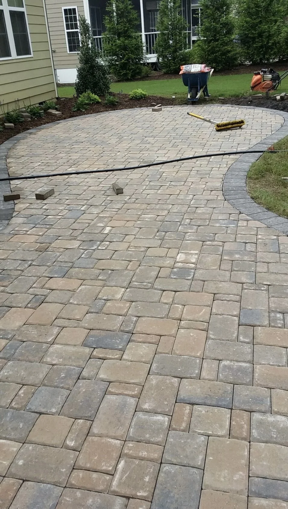 A newly paved stone patio or walkway with construction tools and materials, including a broom, bricks, a hose, and a wheelbarrow, in a backyard with green grass and bushes.