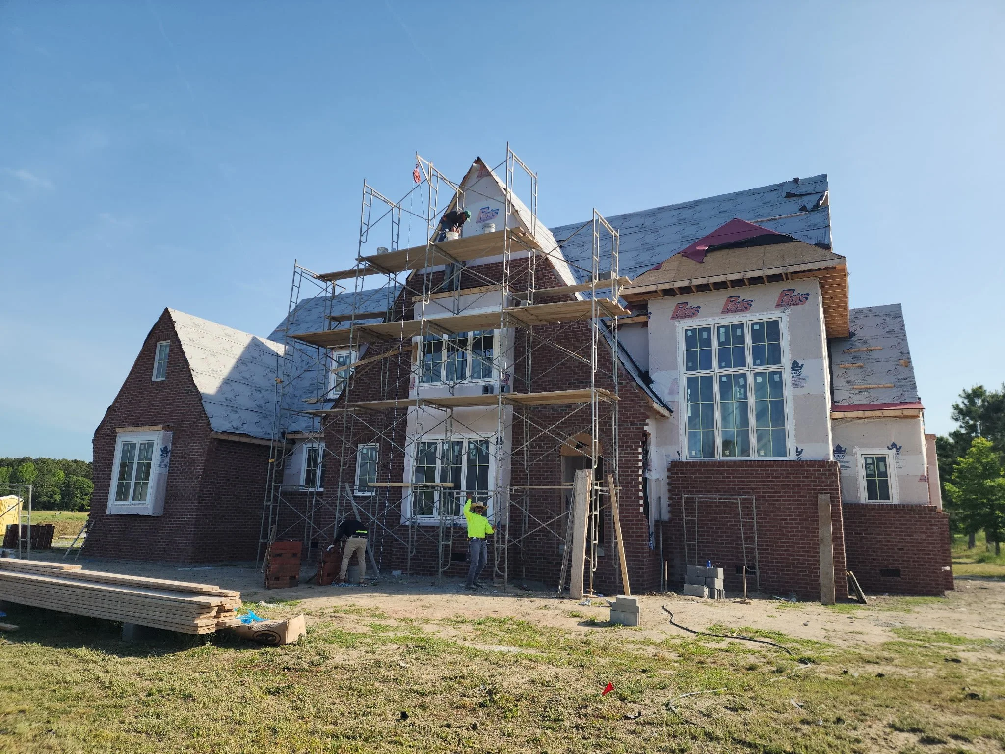 A large house under construction with scaffolding, workers, and building materials in the yard, and a clear blue sky overhead.